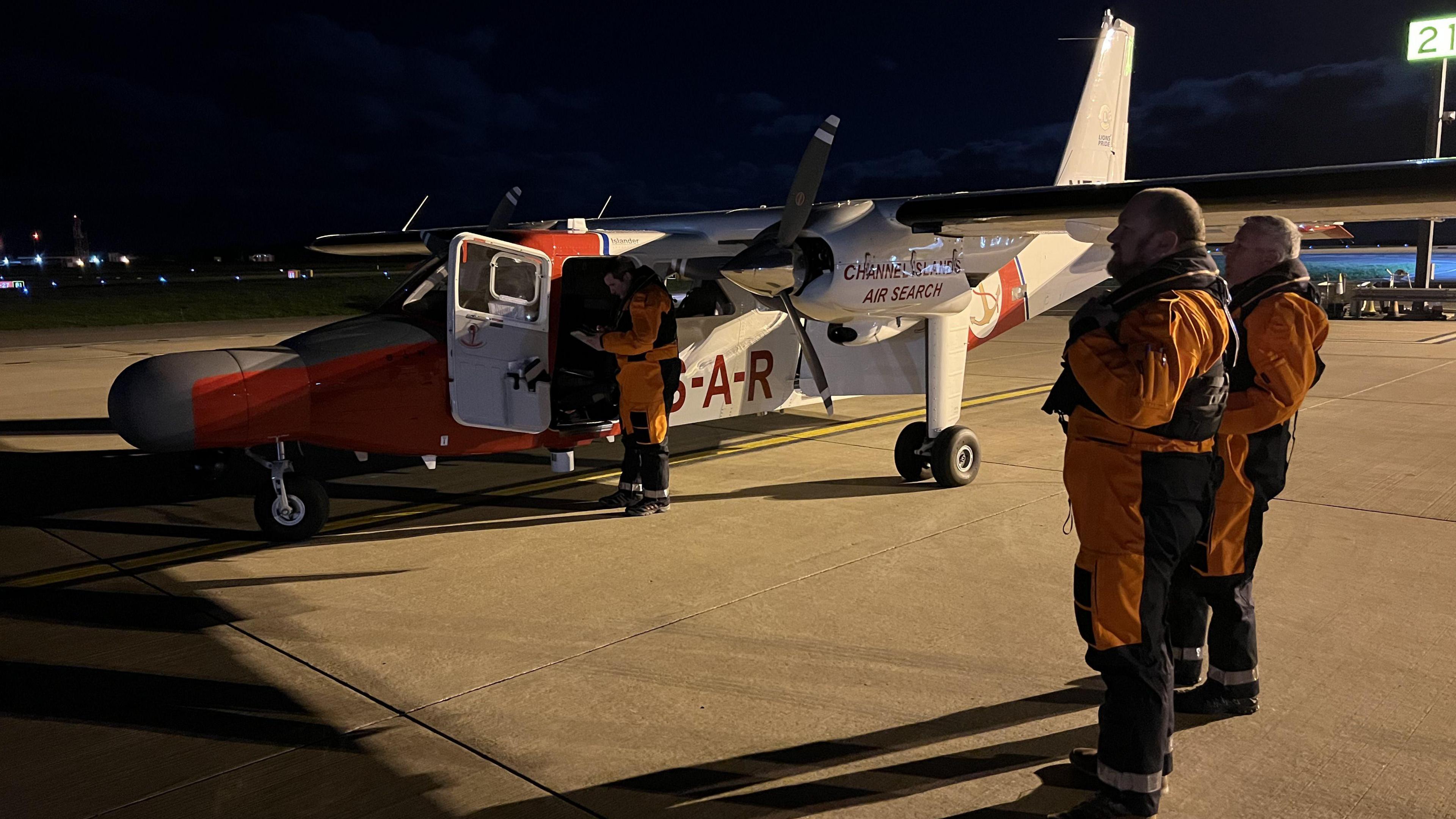 Two crew members from the Channel Islands Air Search team wearing orange overalls on the left. There is another crew member at the door of the aircraft, called Lions’ Pride, which is on the ground. The aircraft is predominantly white with red text and sections.
