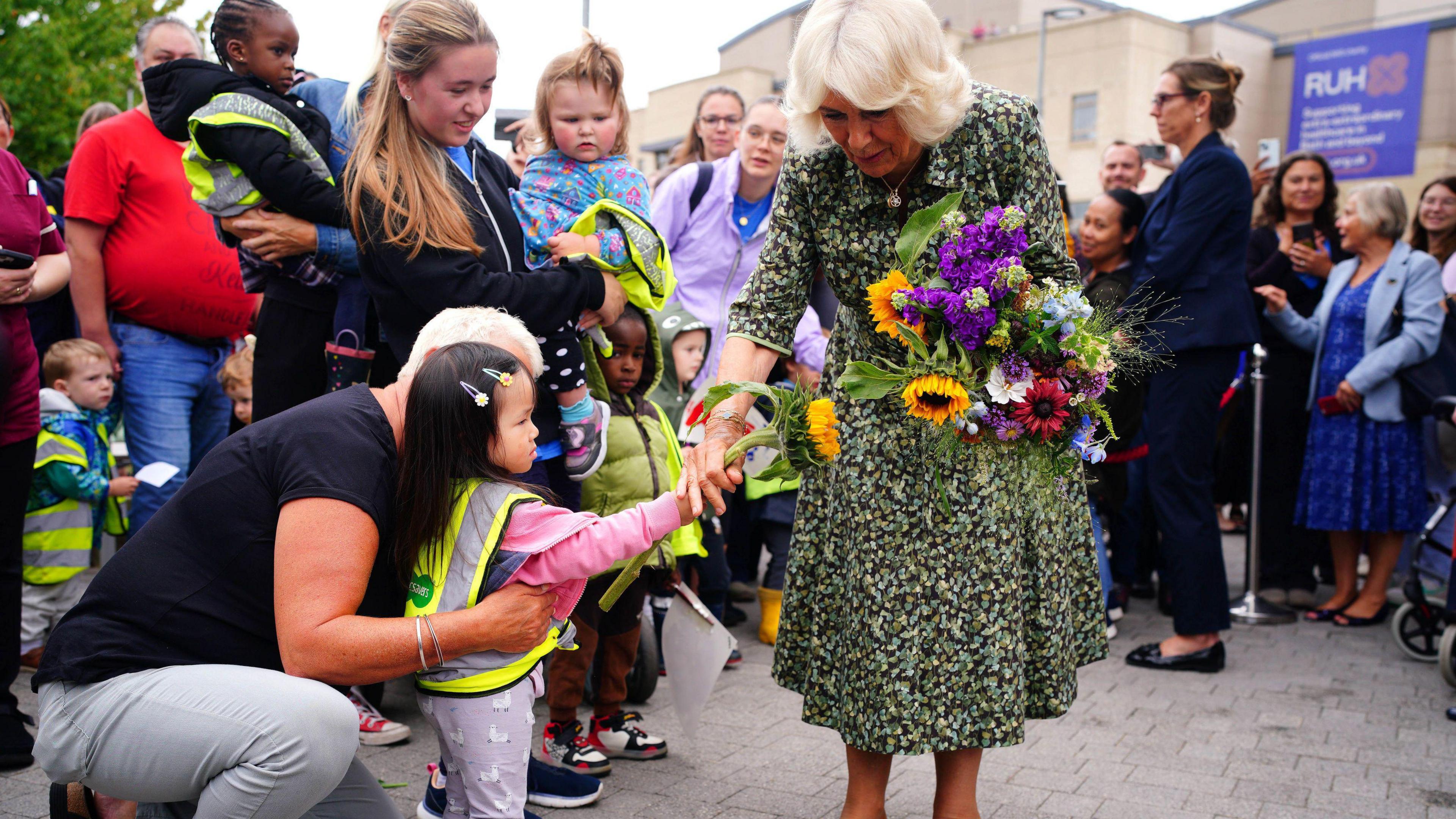 The Queen bending down to receive a sunflower from a child during her visit to the Dyson Cancer Centre