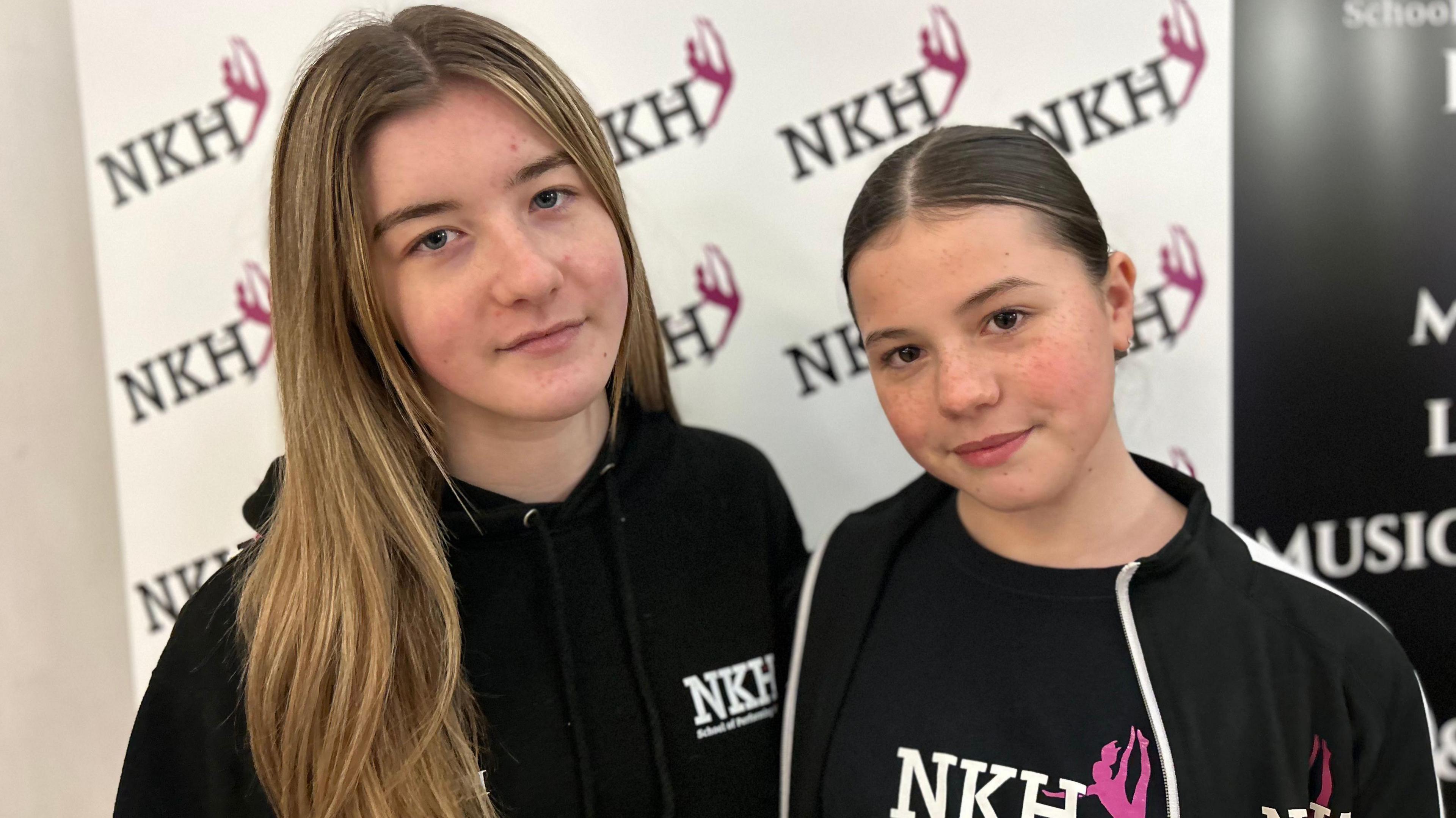 Two young girls looking at the camera with glum faces wearing their dance company's merchandise. Their clothing is black with white writing, and they're standing in front of a board with their dance school's logo on.