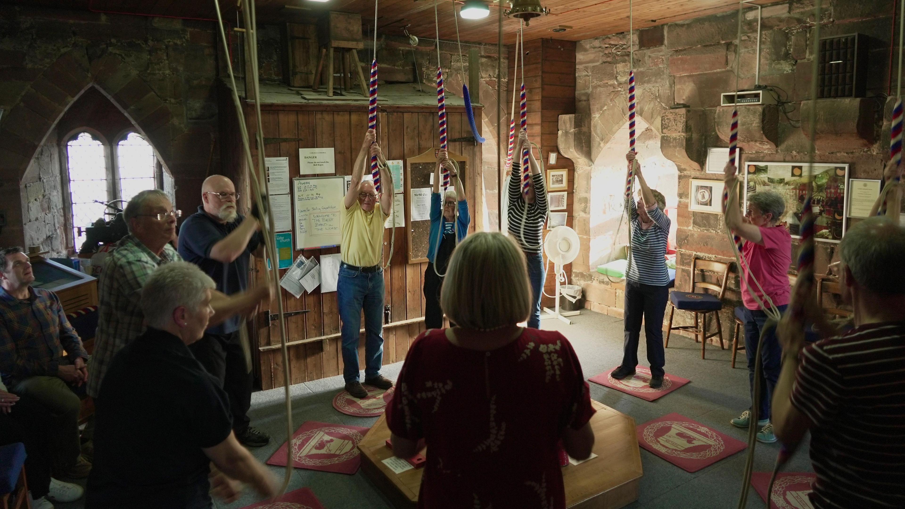 A group of men and women in a church bell tower, reaching up to large ropes hanging down from the ceiling. It is brightly lit from church windows.