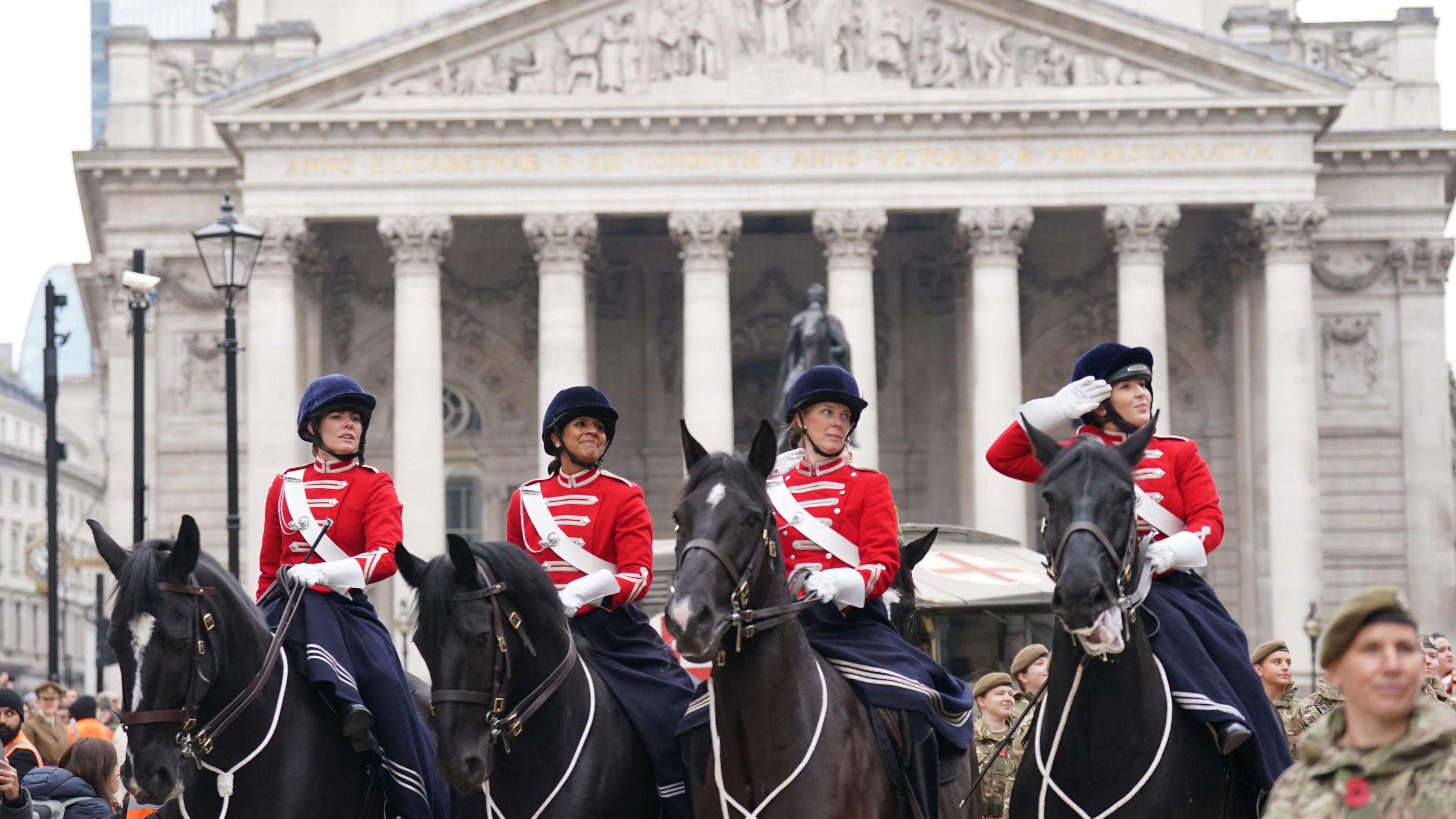Four female horse riders take part in the Lord Mayor's Show in the City of London in November 2024. They are each wearing bright red military-style jackets with white sashes along with dark blue hats. They are in front of the official residence of the Lord Mayor of London. It is a grand building in the Palladian architecture style.