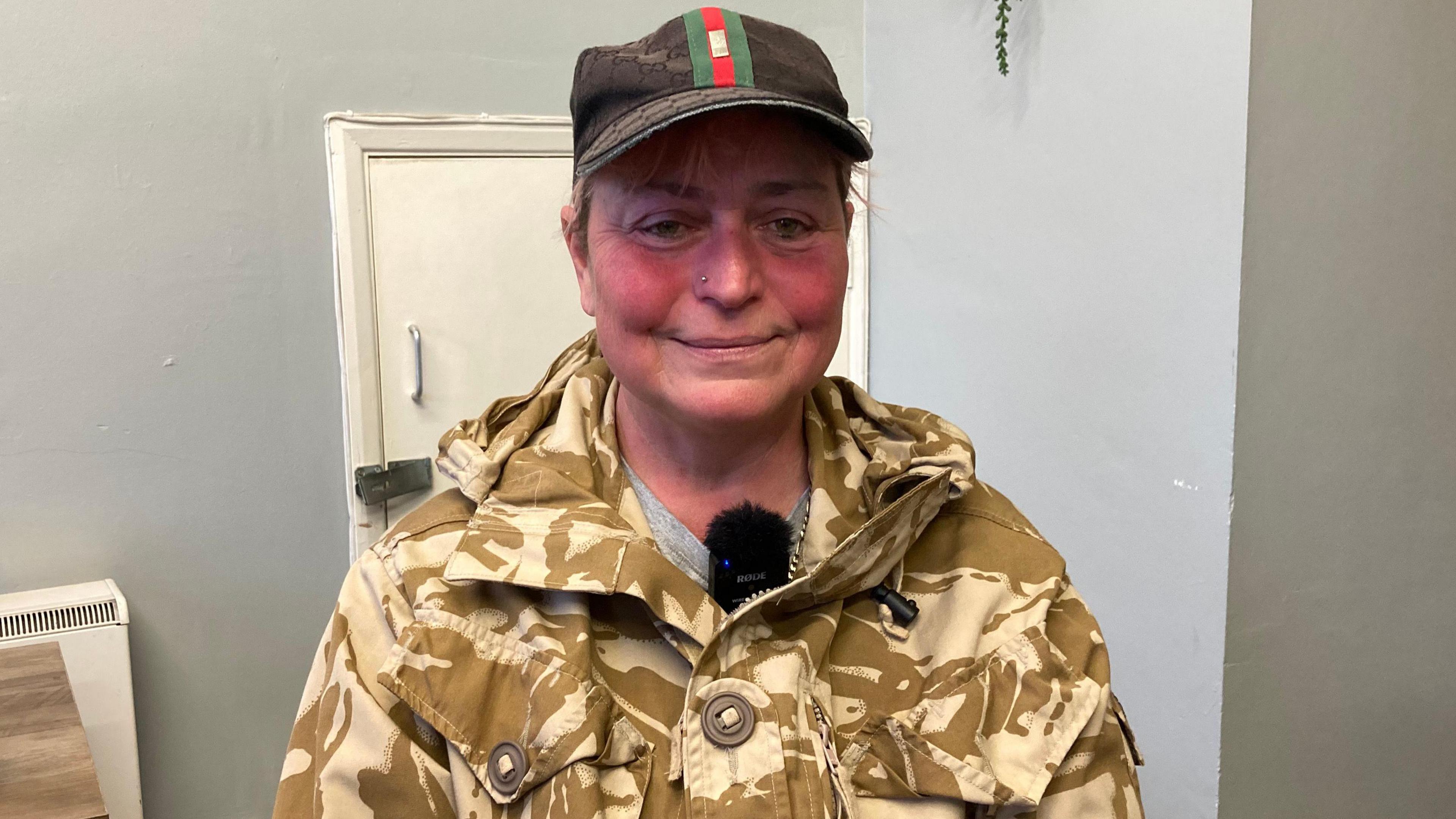 Emma Goodridge. She is wearing a black hat with a green and red stripe on it. It is covering her hair. She has a nose stud and is wearing a camouflage jacket jacket, looking directly at the camera and smiling.  