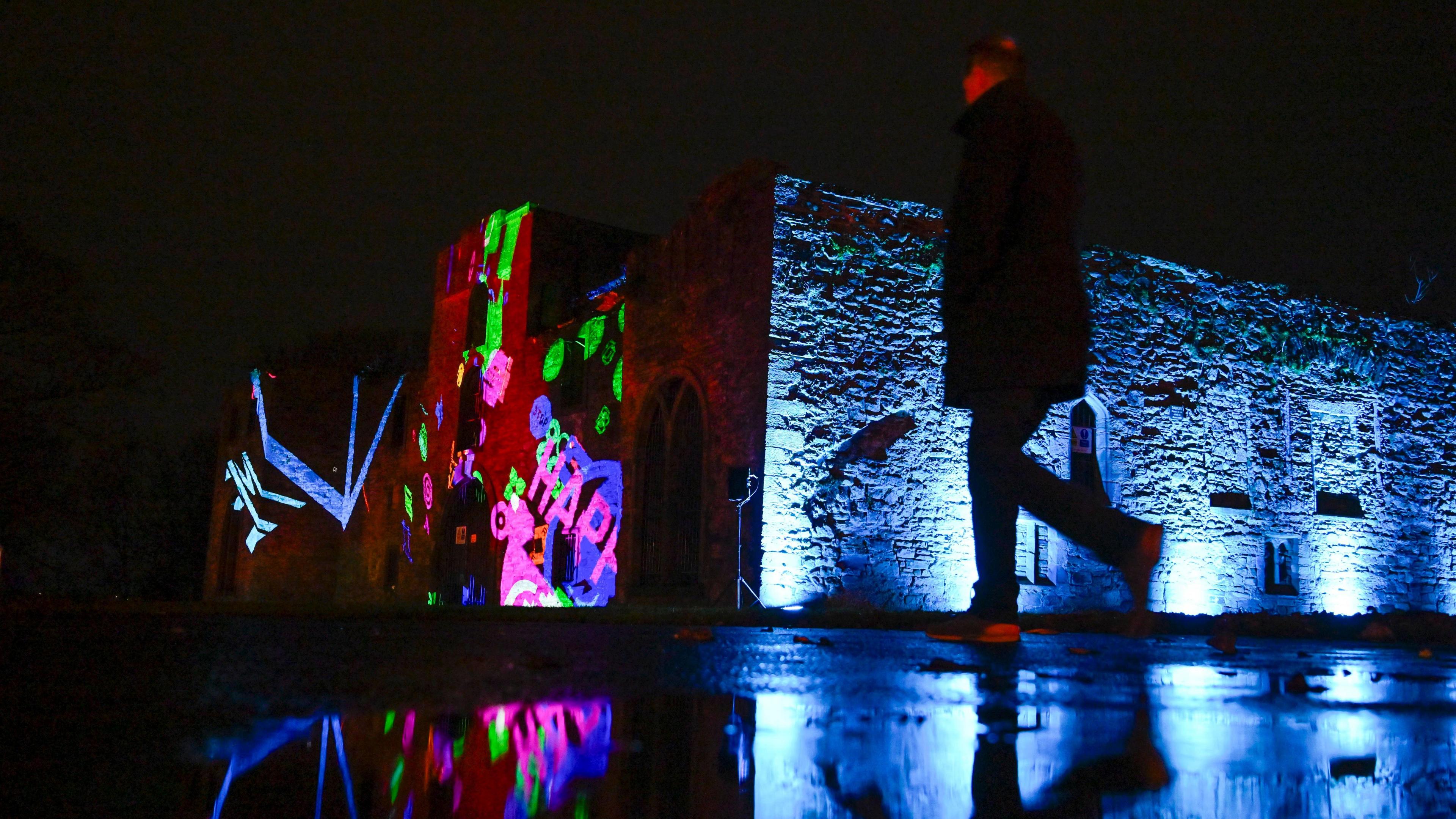 Designs of various colourful symbols are projected onto Workington Hall. A person is walking in front.