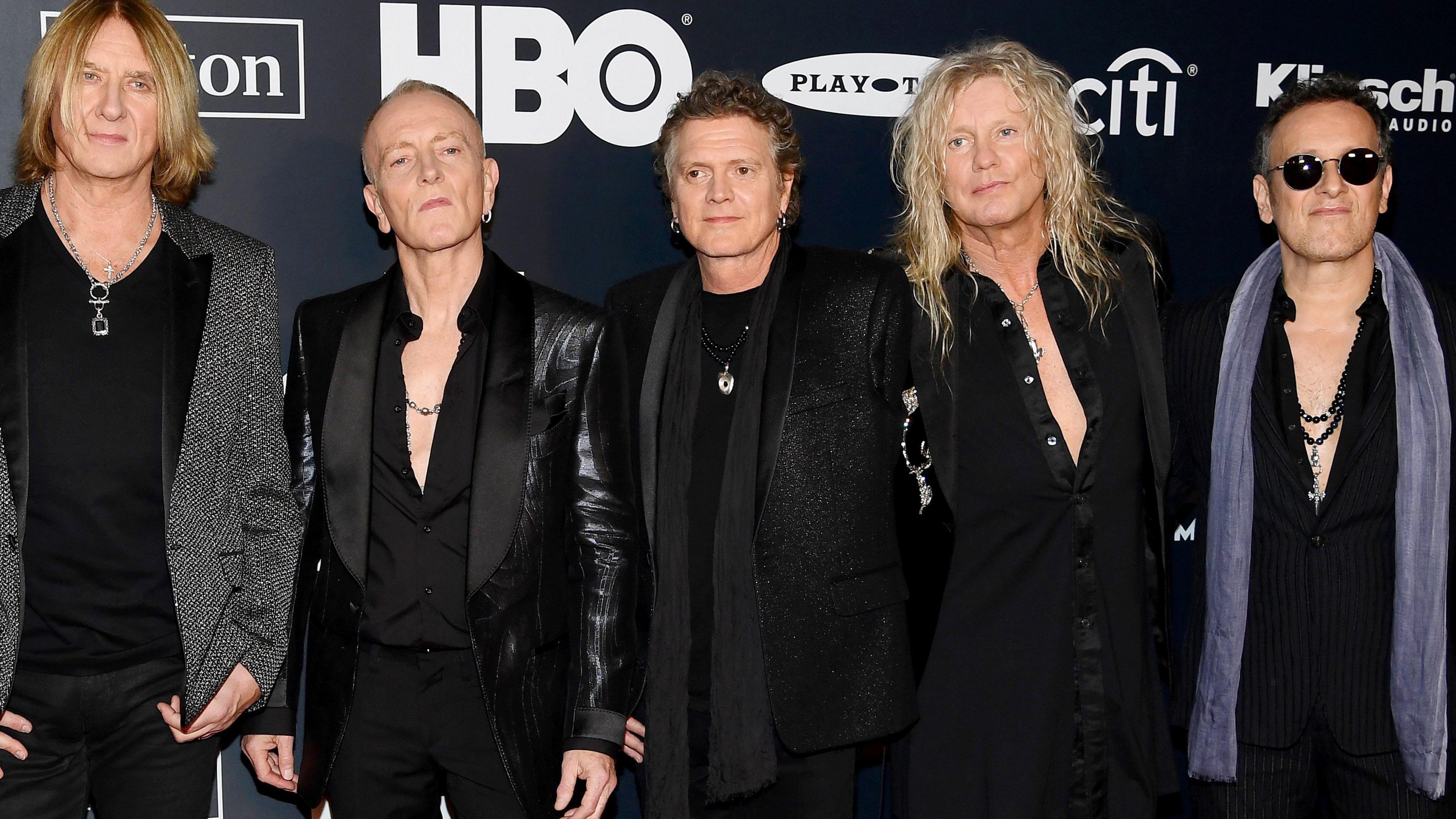 Joe Elliott, Phil Collen, Rick Allen, Rick Savage and Vivian Campbell of Def Leppard attend the 2019 Rock & Roll Hall Of Fame Induction Ceremony. There are logos behind them on the backdrop. The men are dressed in black and Vivian Campbell has a grey scarf on.