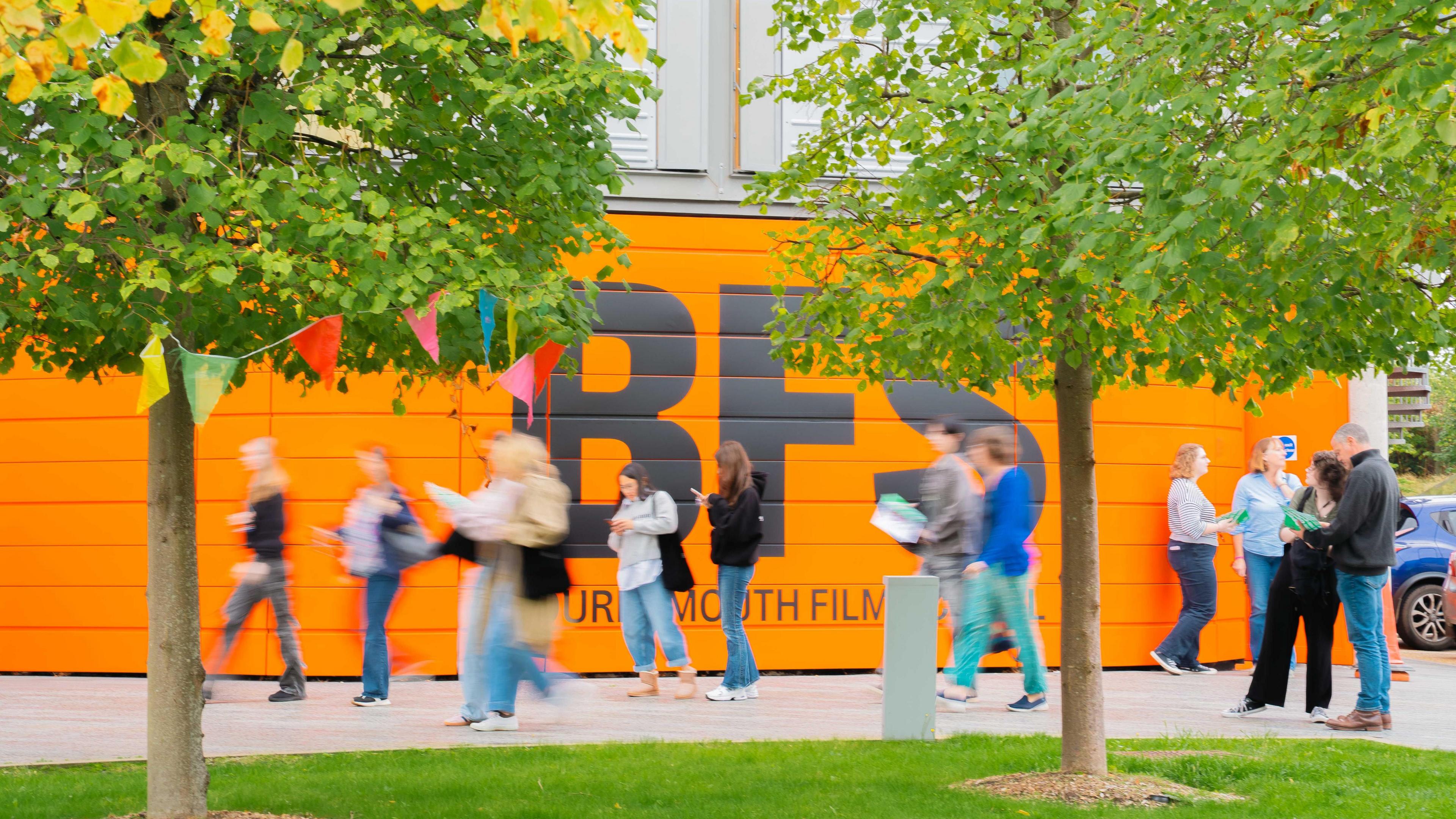 Students gather and walk past the outside of the film school, which has a bright orange exterior wall with BFS across it in black letters.