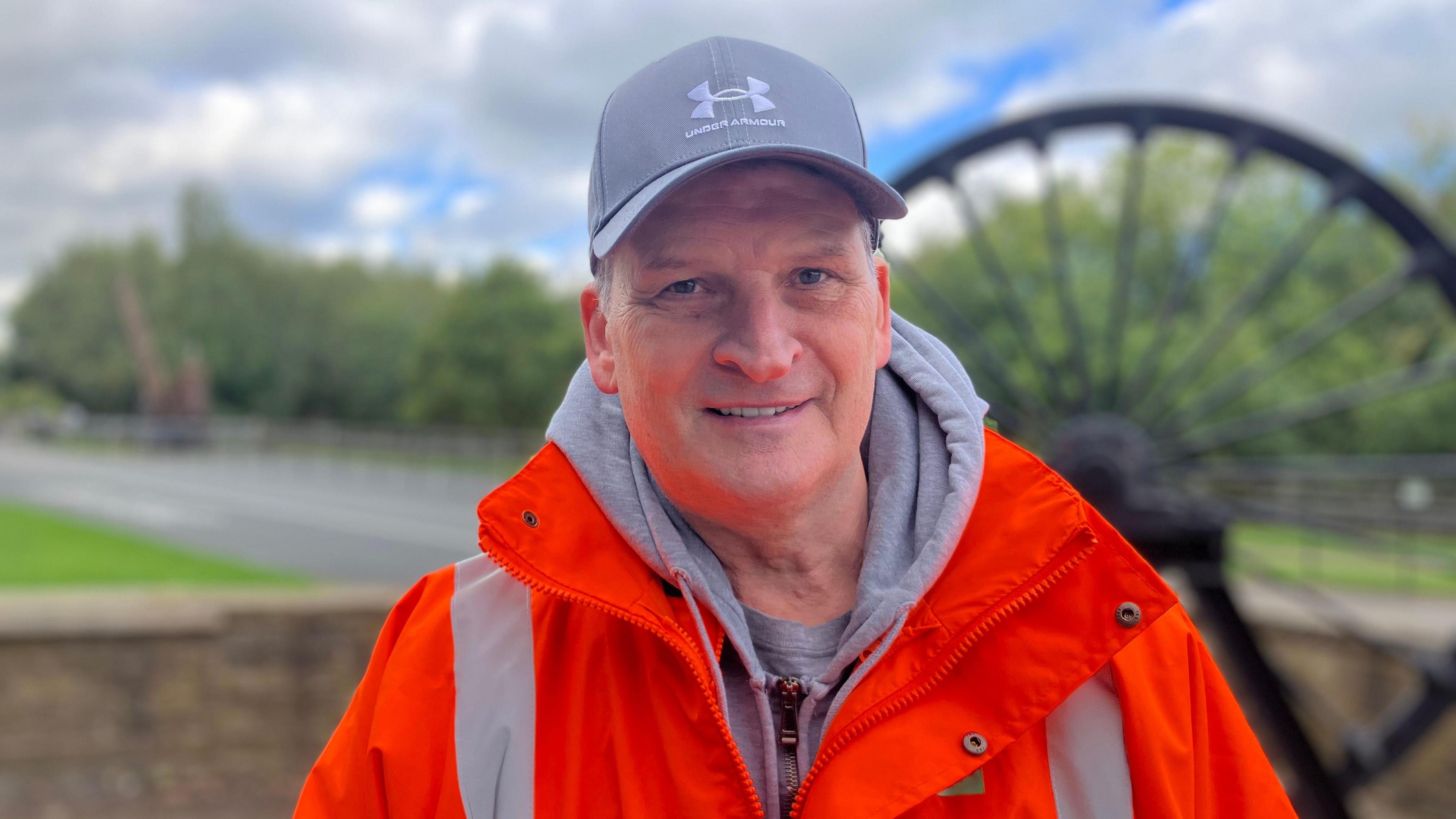 A man in an orange high vis smiles at the camera. He wears a grey cap and  grey hoodie. Behind him, a water wheel is blurred.