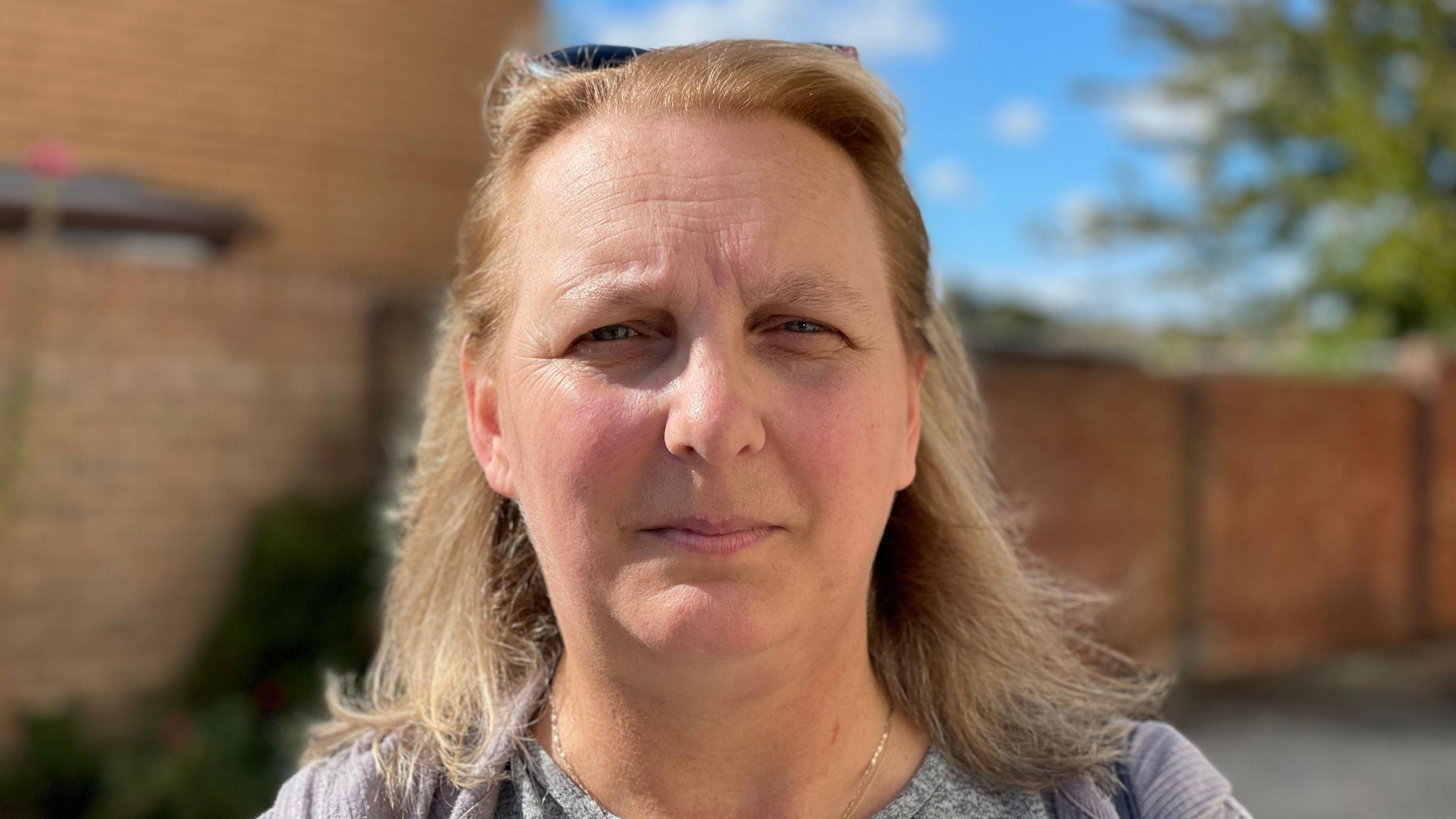 Jo Woods is looking straight at the camera. She has shoulder-length fair hair and has sunglasses balanced on the top of her head. She is wearing a grey top and has a chain around her neck