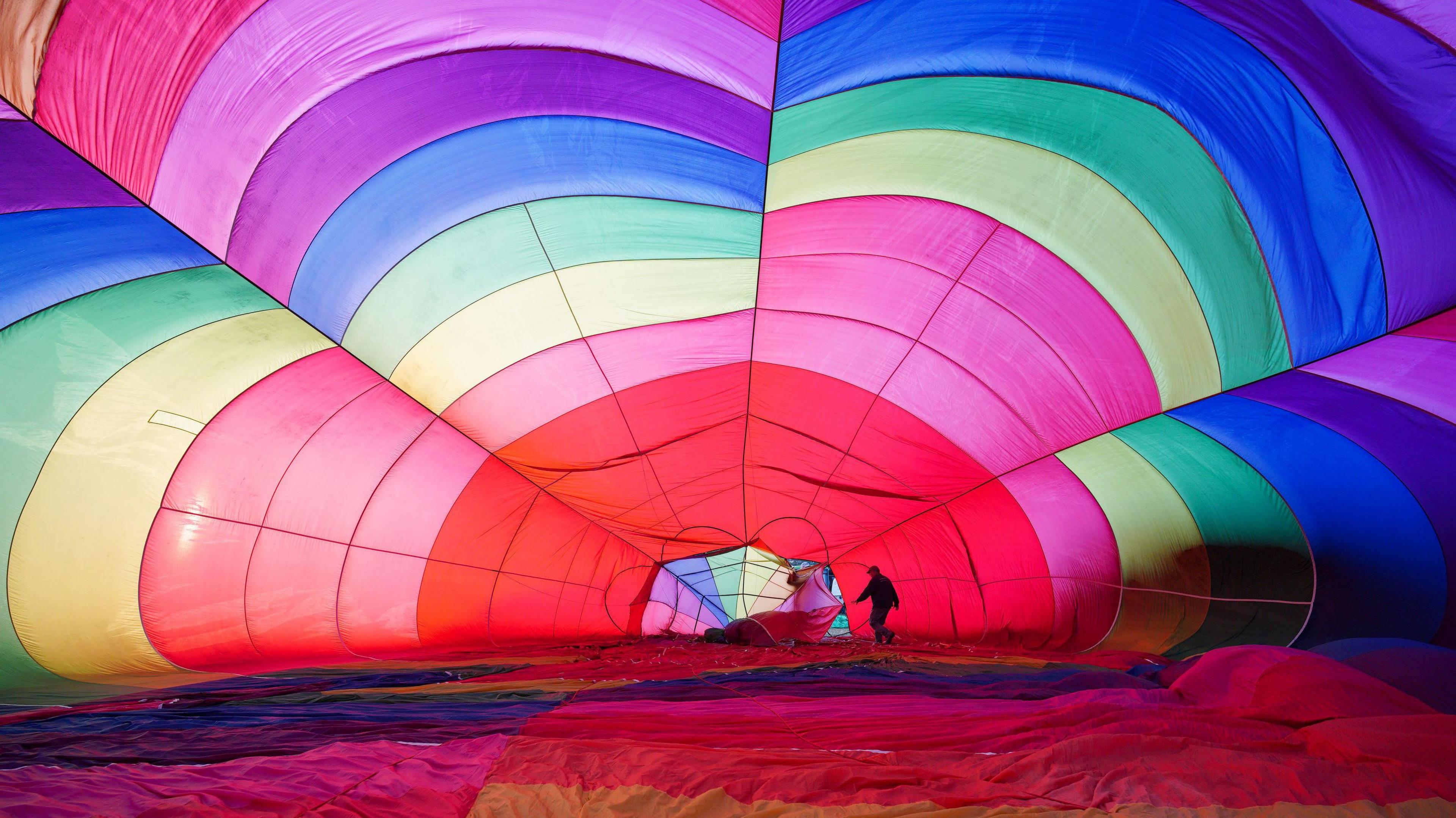 The inside of a hot air balloon, which is all colours of the rainbow, as it is being inflated. A man can be seen walking inside it, towards the opening where the basket normally goes.