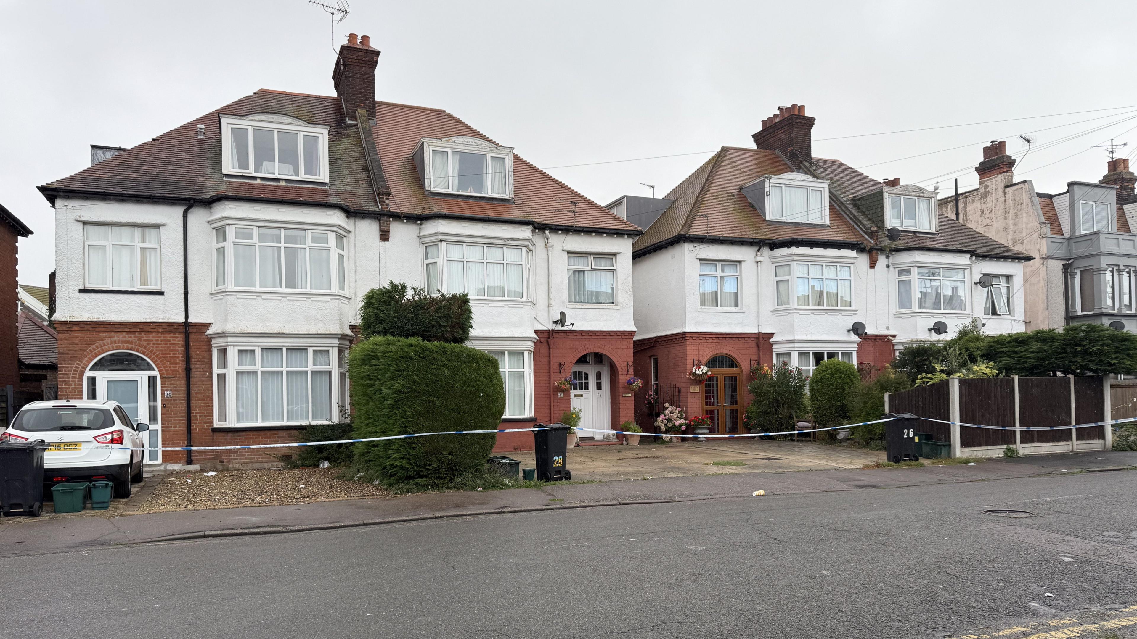 Four semi-detached houses behind a police cordon. There is a grey cloudy sky.