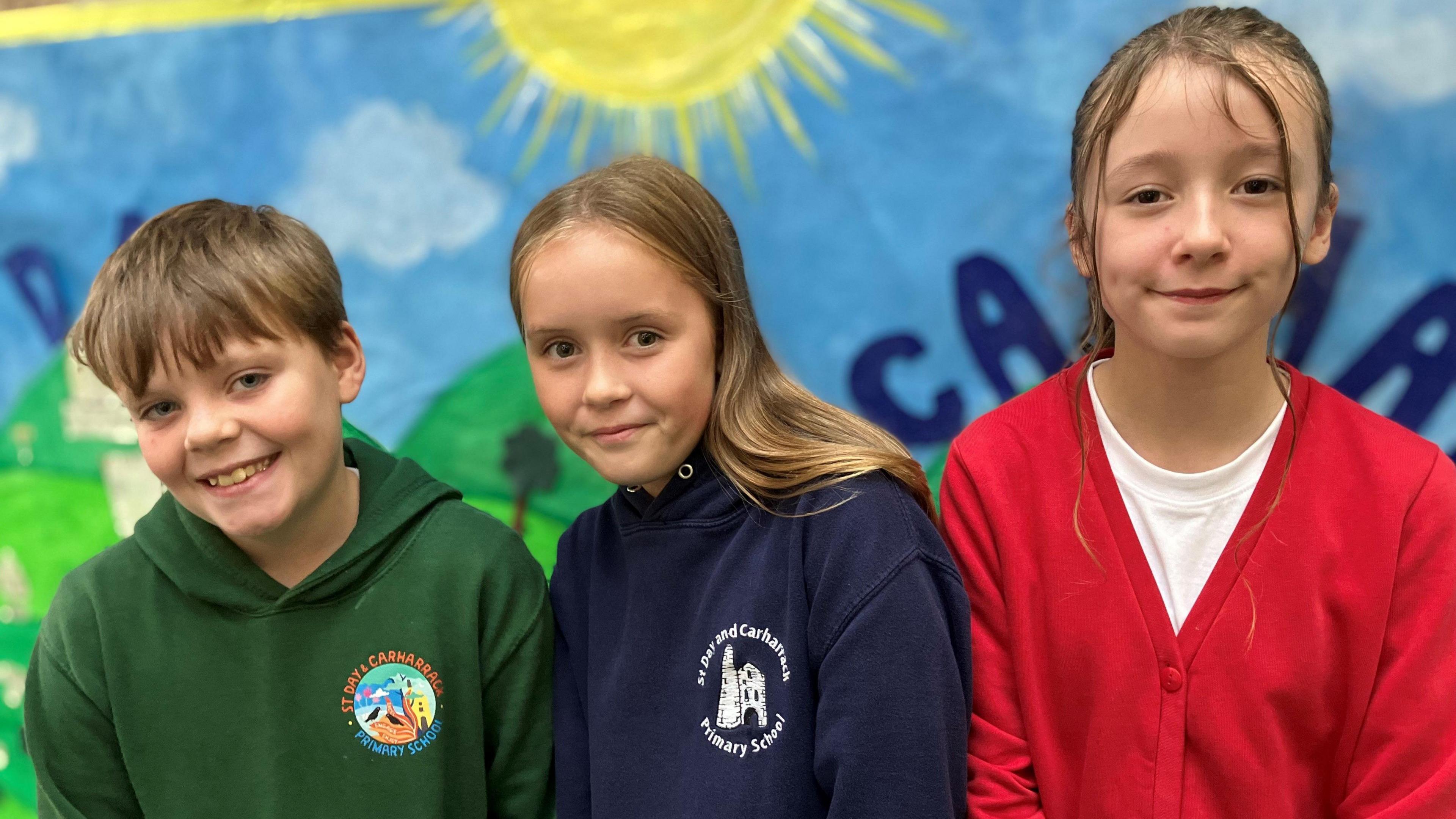 Three pupils smiling at the camera. There is a boy and two girls standing in front of a big mural with the sun on it and blue skies.