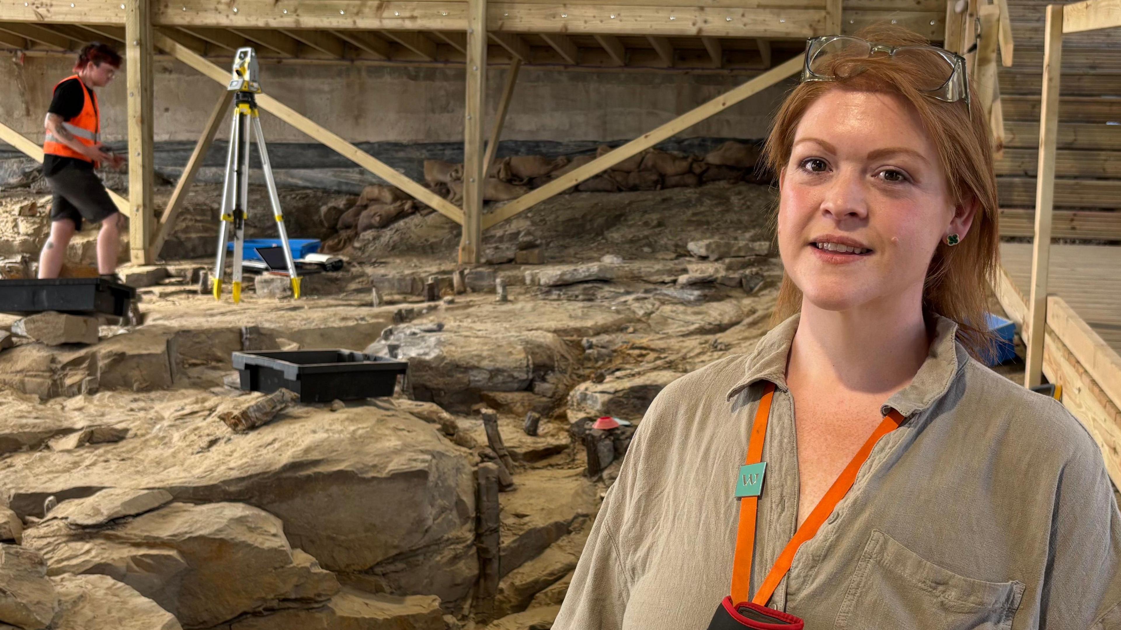 Amy Rattenbury smiling at the camera standing on part of the rocky fossil forest site with a student in the background using equipment to study it. She has long red hair and has glasses perched on top of her head, and wears a brown shirt.