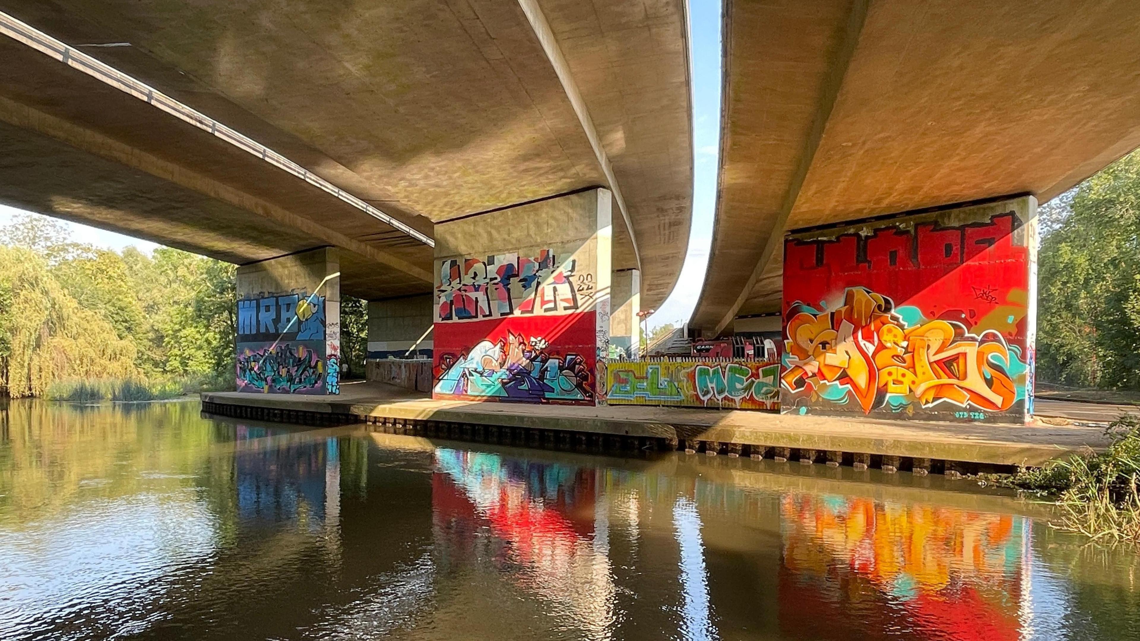 A striking image of a mural painted onto the walls of an underpass. The graffiti is bright red, yellow and blue on the uprights of a bridge that appears to be carrying a motorway over a river. The bridge and pictures are reflected in the water. Either side of the bridge you can see trees and sunshine is streaming through the gaps where you can see glimpses of blue sky. 