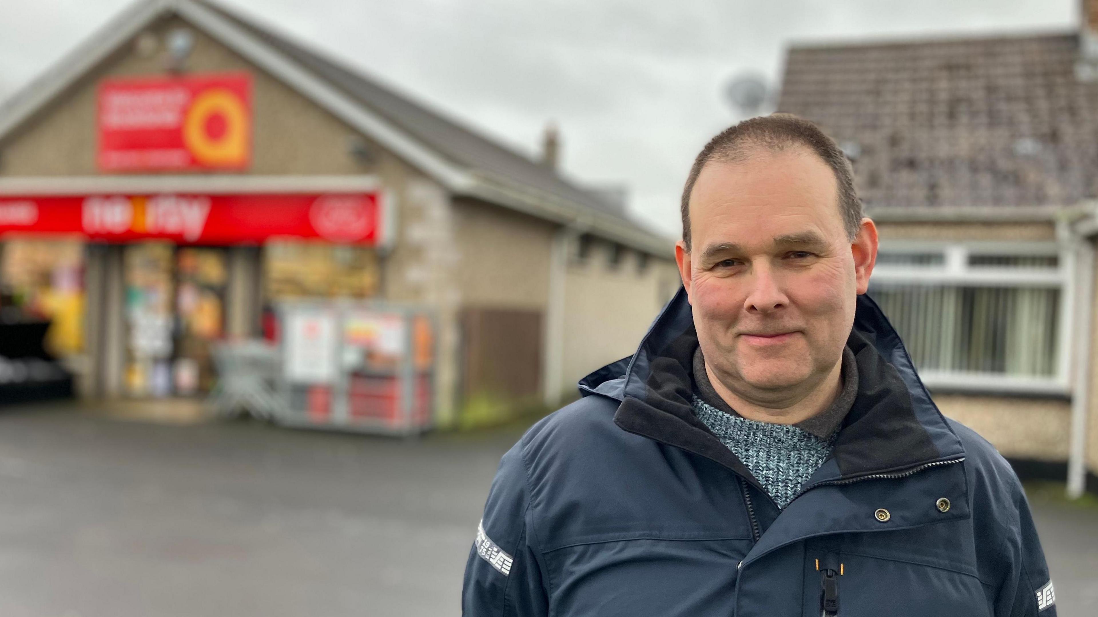 Gareth Pidgeon - he has short dark hair and is wearing a navy blue coat over the top of a light blue cable-knit sweater. In the background his shop can be seen, it is a light coloured building with a red sign.