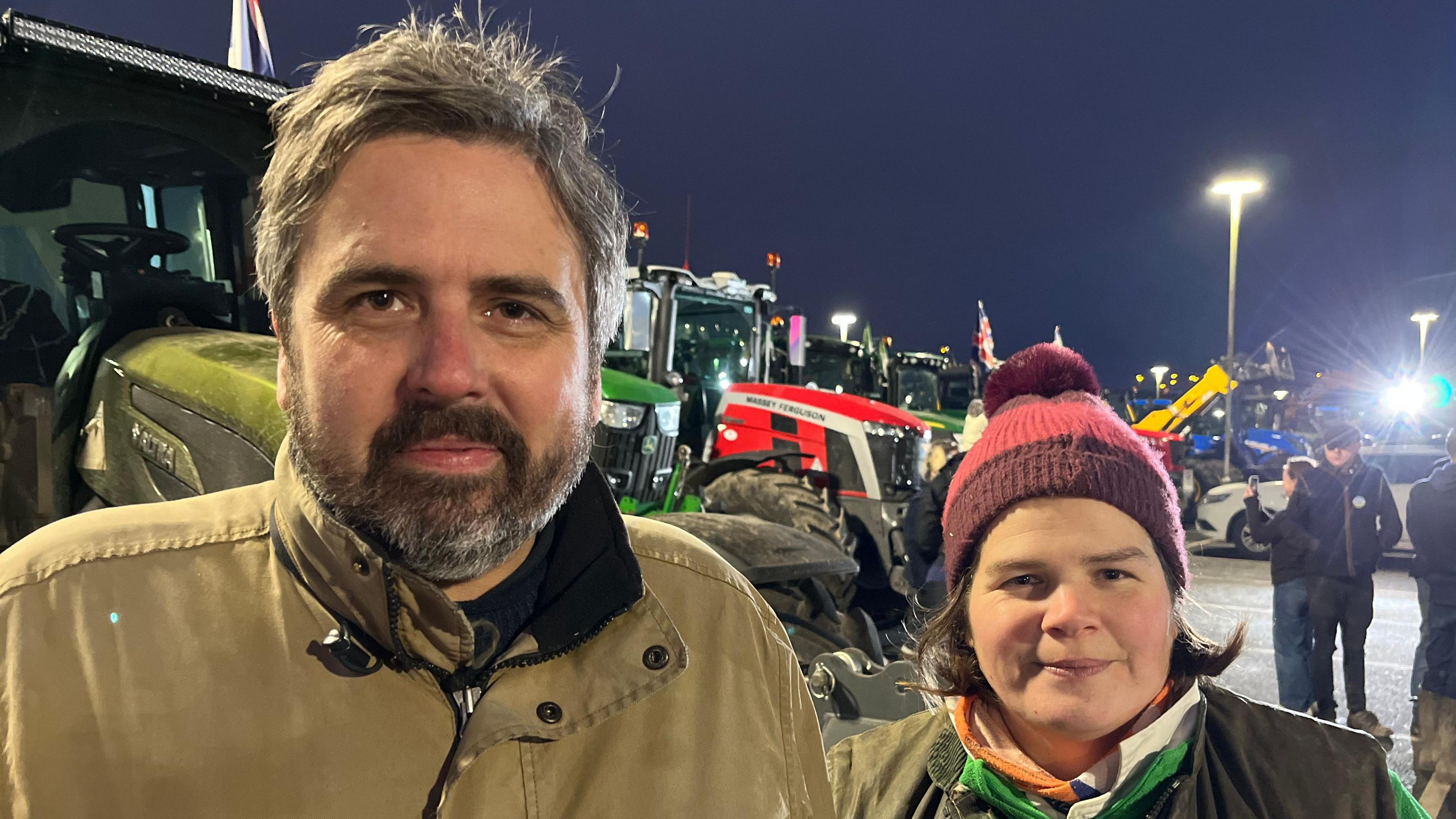 A bearded man in a brown jack and a woman with chin-length brown hair wearing a hat and scarf stand in front of a row of tractors. 