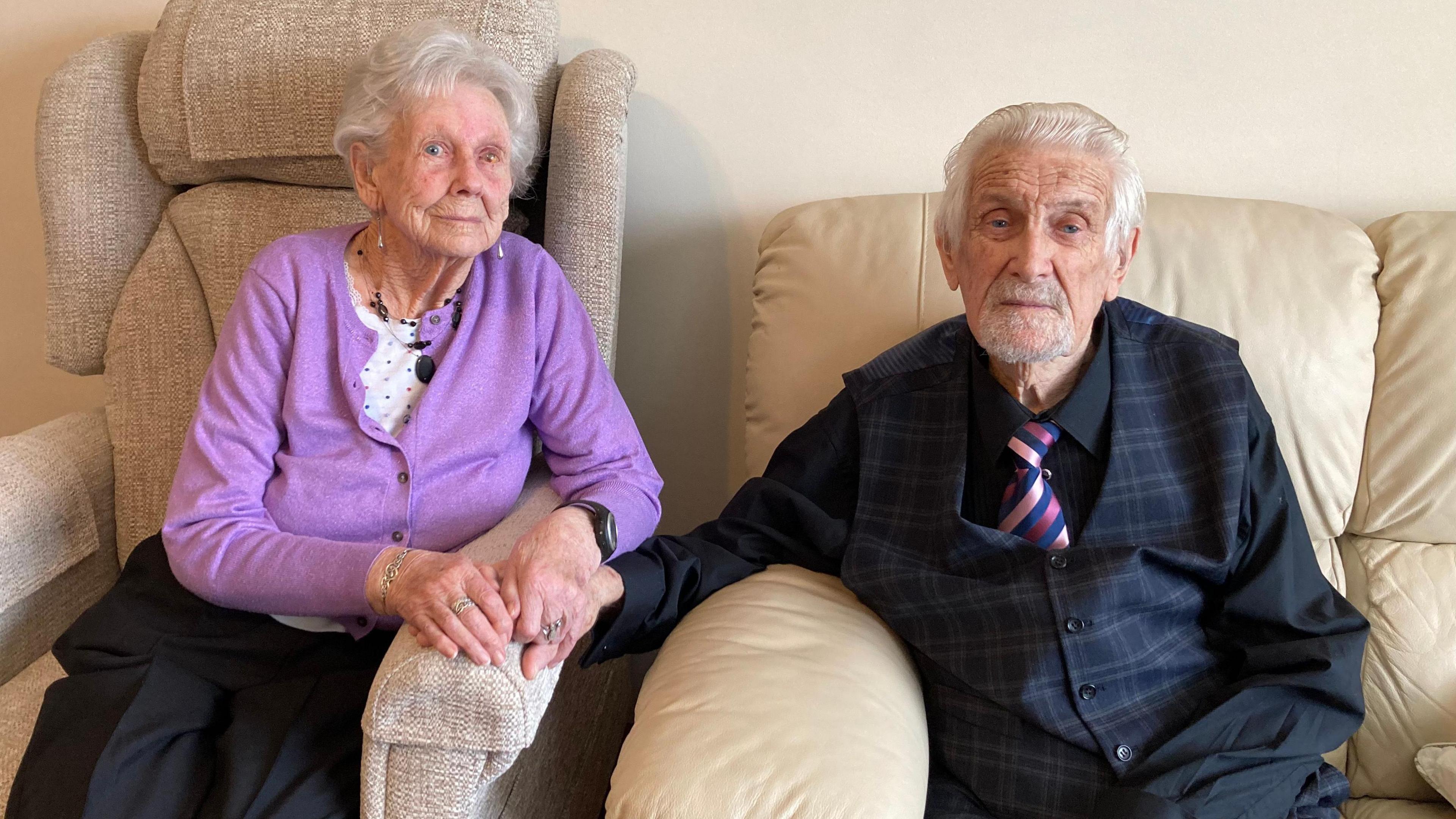 Derek (right) and Brenda (left) Dodge sitting on two chairs side-by-side, holding hands in their home on their 70th wedding anniversary.