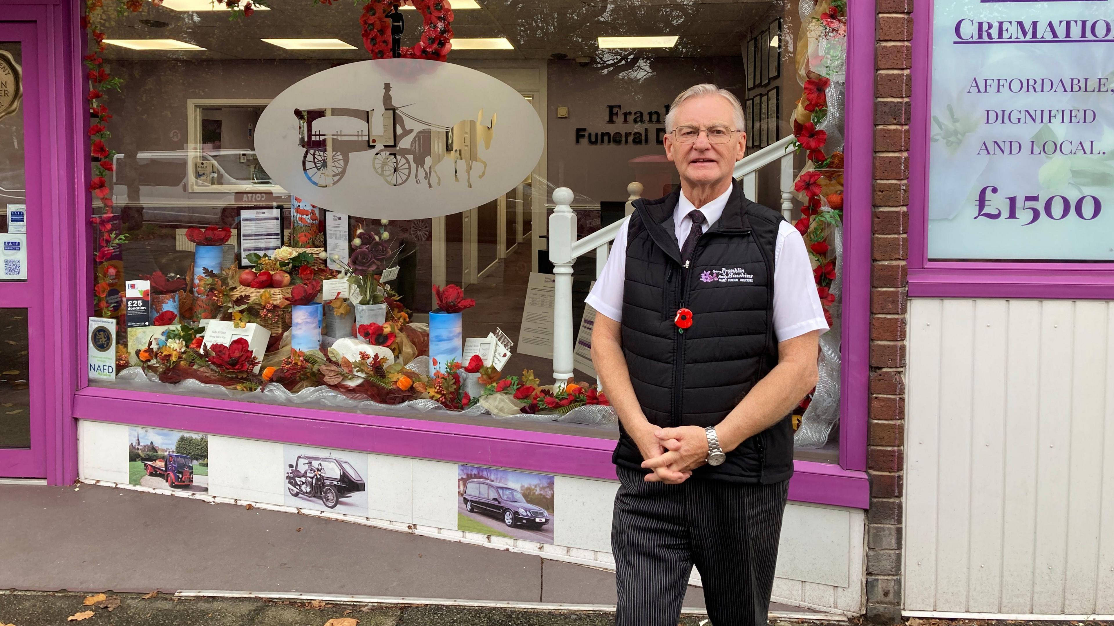 A man with short grey hair standing in front of a funeral director's premises. The building's window frames are painted purple, with a seasonal window display featuring poppies and autumn leaves.
