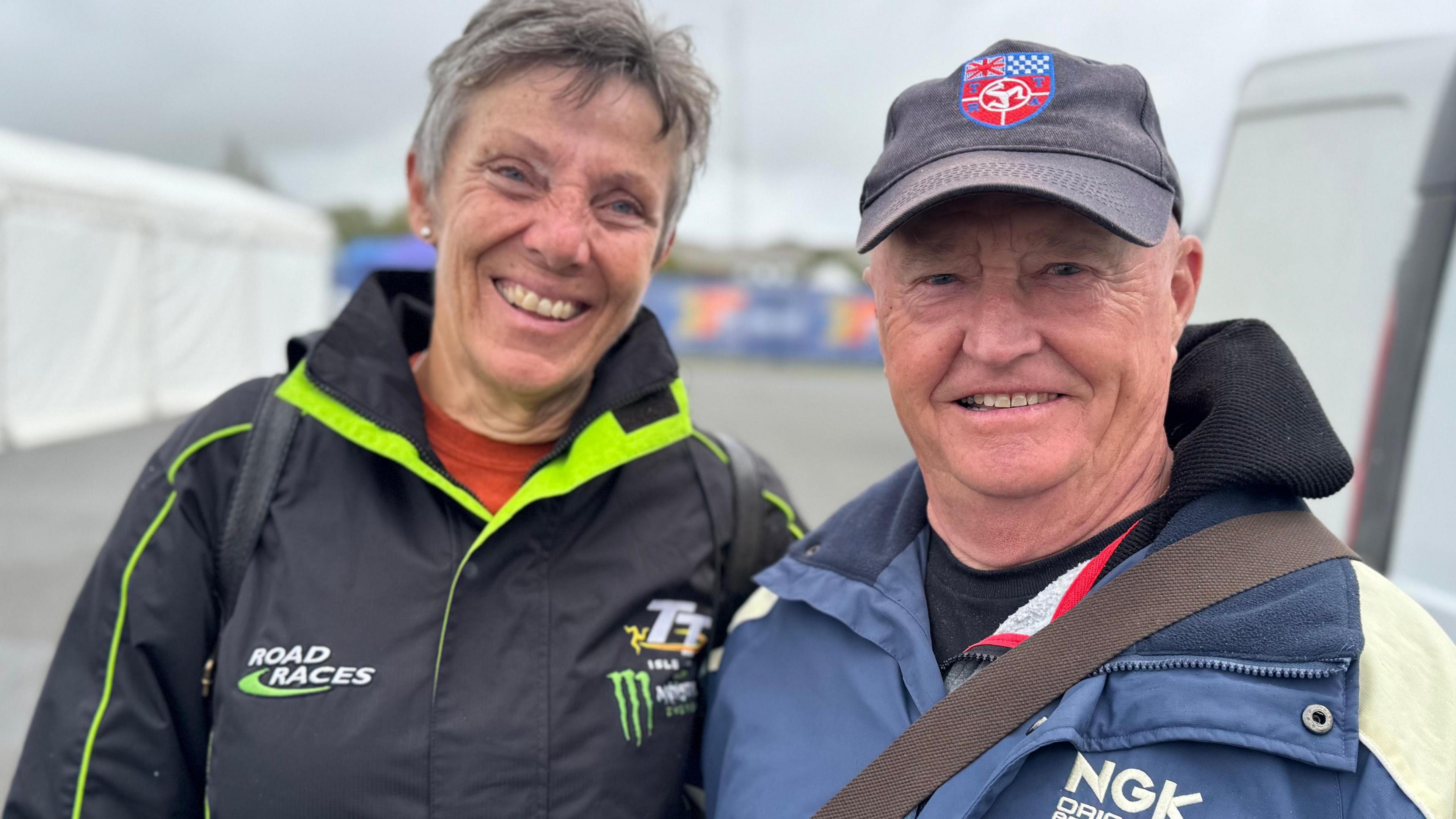 A man and woman smile next to a white tent and white van. He wears a blue and cream jacket and a blue cap. She wears a black coat with green trim and a TT logo. She has short grey hair.