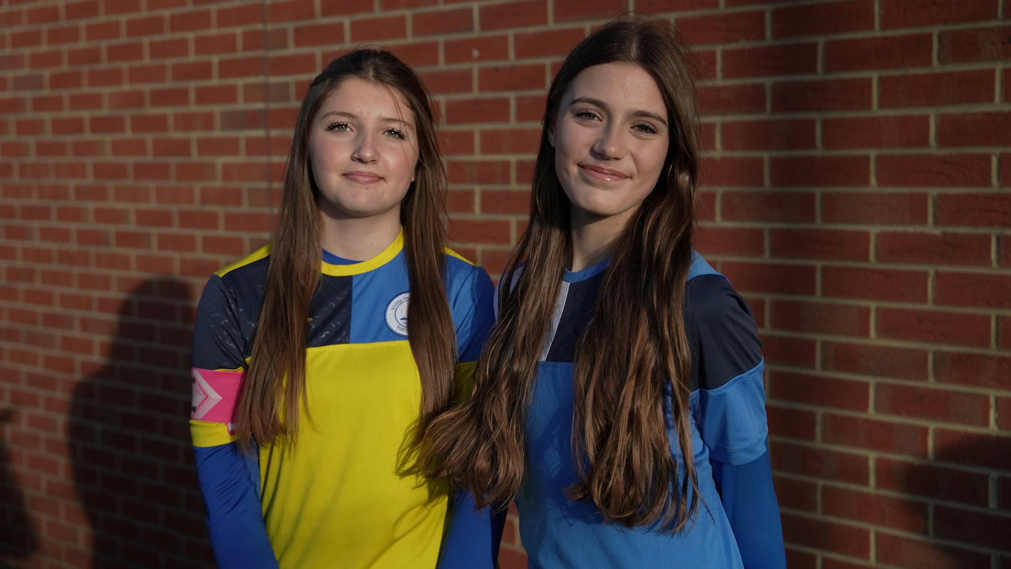 Tiffany and Lana in football kit. They both have long brown hair and are smiling. They stand in front of a brick structure. 