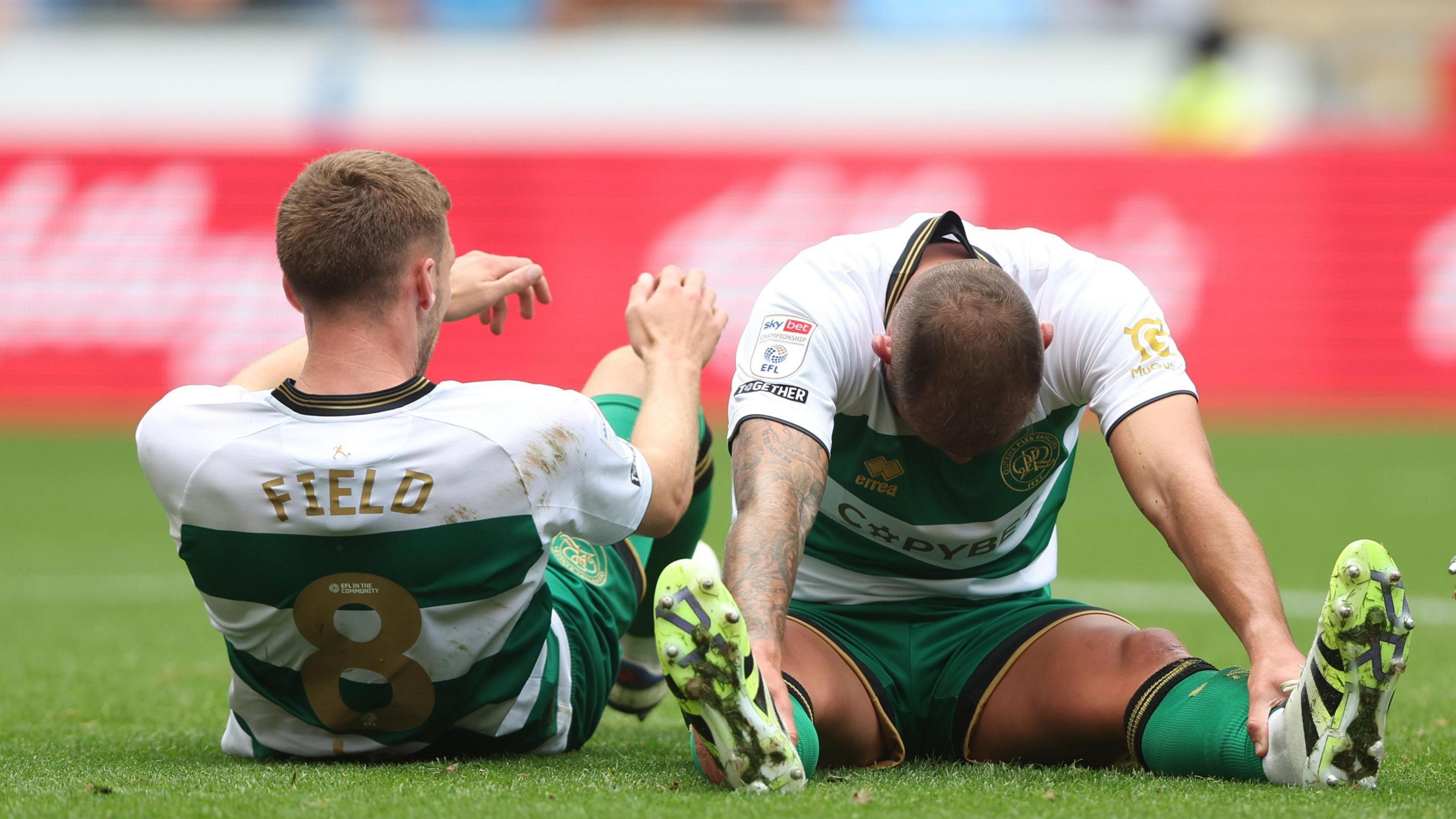 Two QPR players sit side by side on the pitch after full-time as one holds his head forward looking down and stretches his arms to hold his ankles.