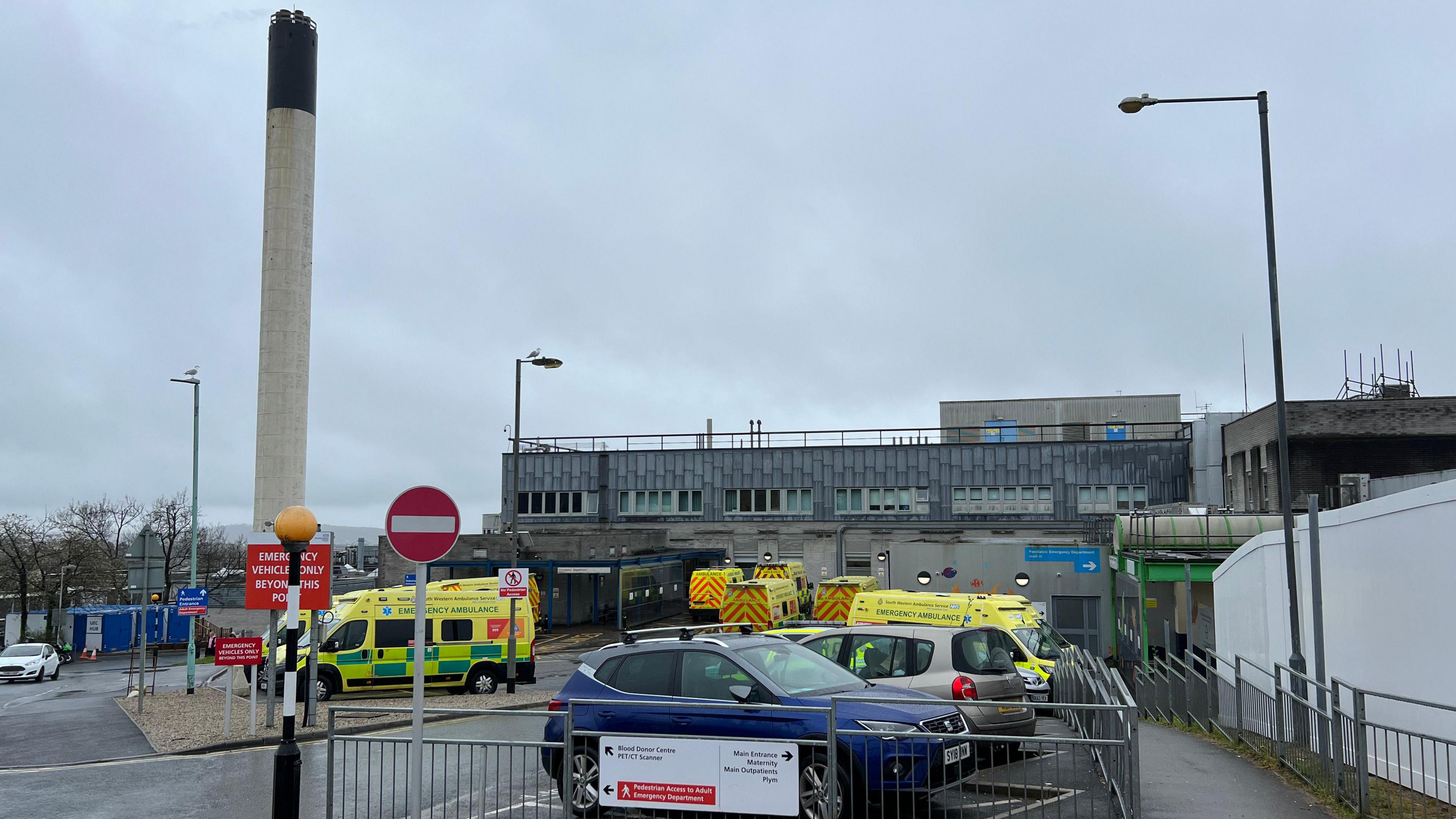 Several ambulances and a couple of cars parked outside Derriford Hospital in Plymouth on a gloomy day. A large chimney is in the background.