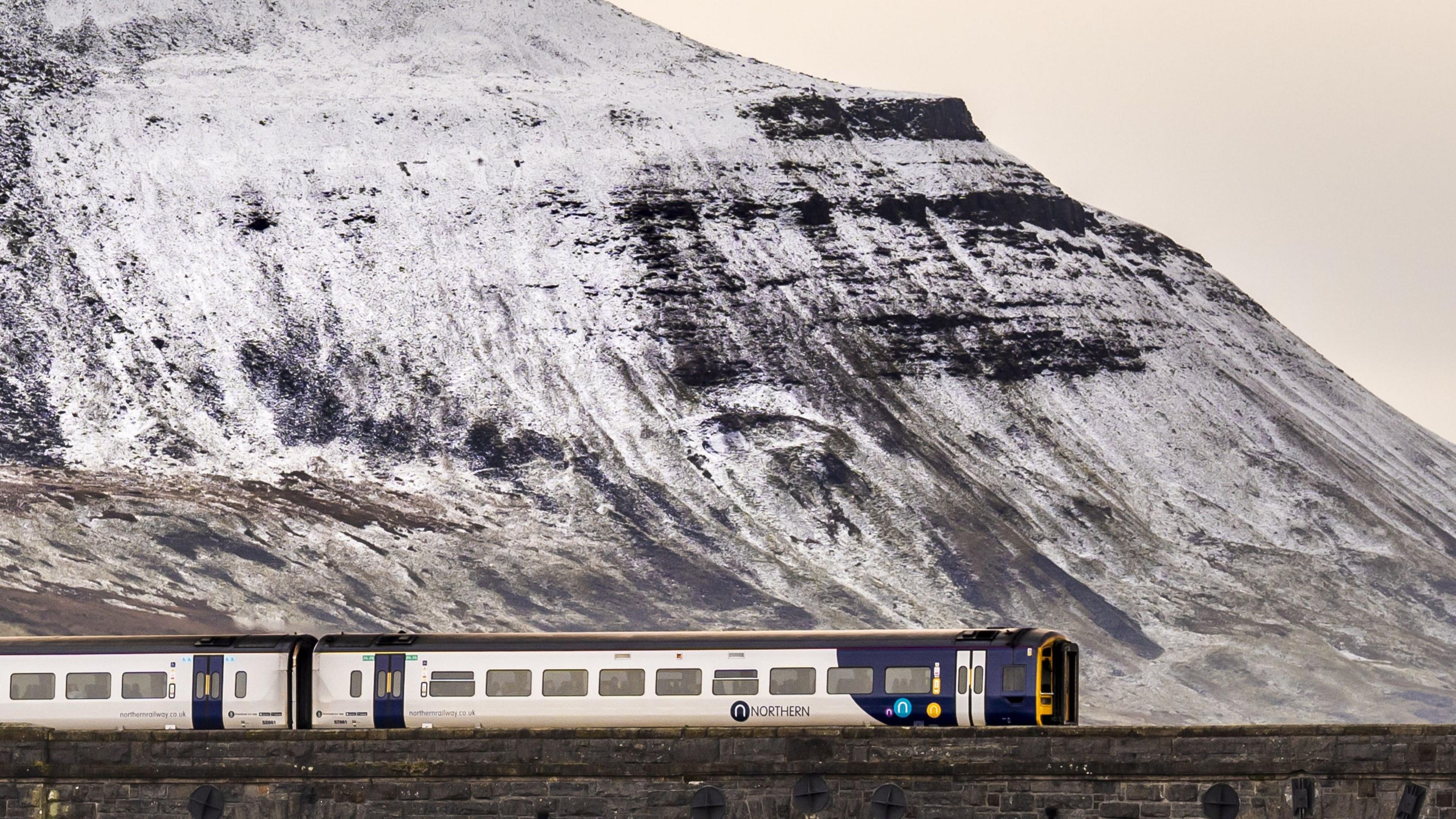 A Northern train travels over a stone bridge with snow on a rocky hill in the background in the Yorkshire Dales