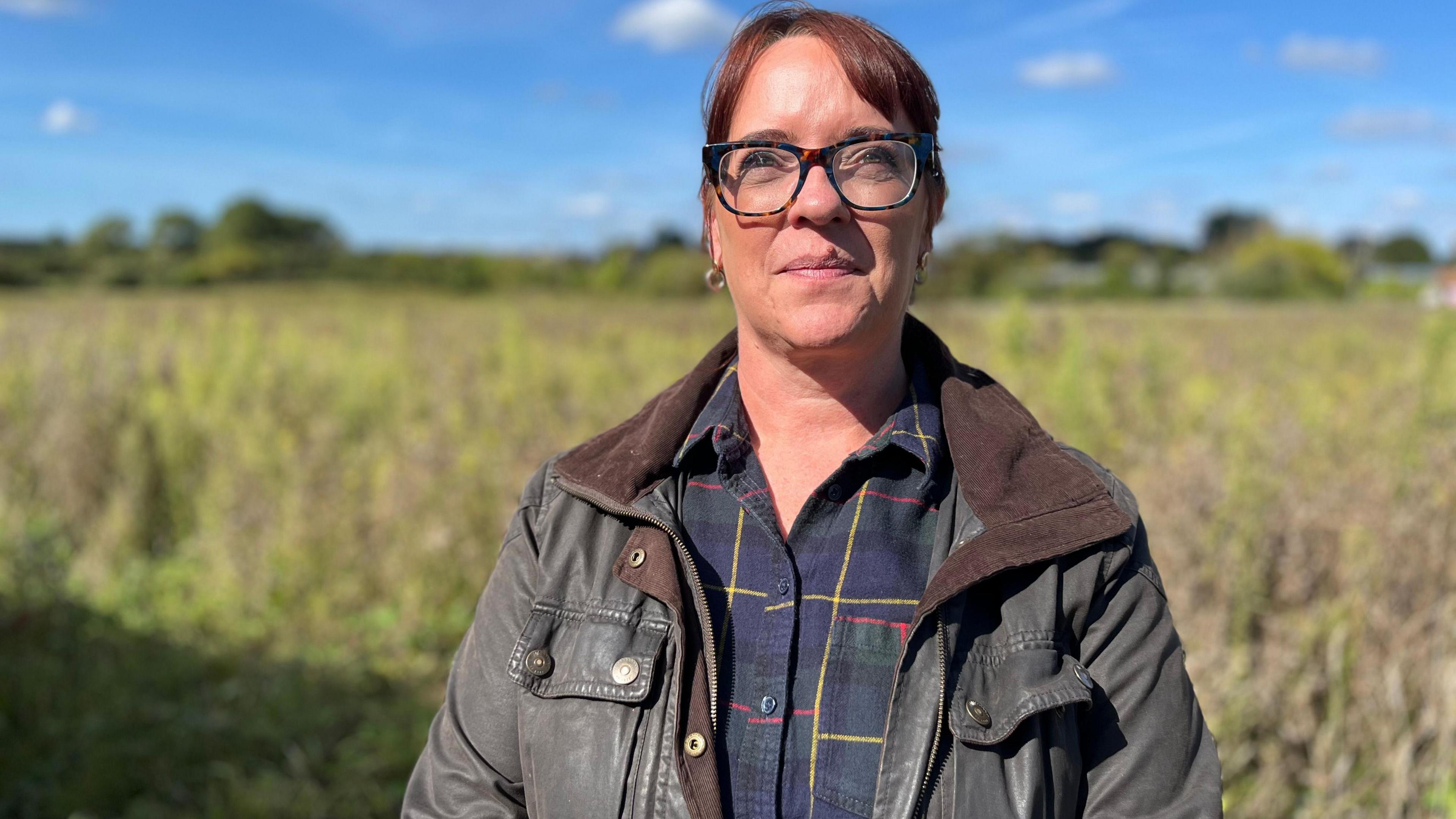 A woman with a green Barbour jacket and a checked shirt, with black glasses looking towards the camera.