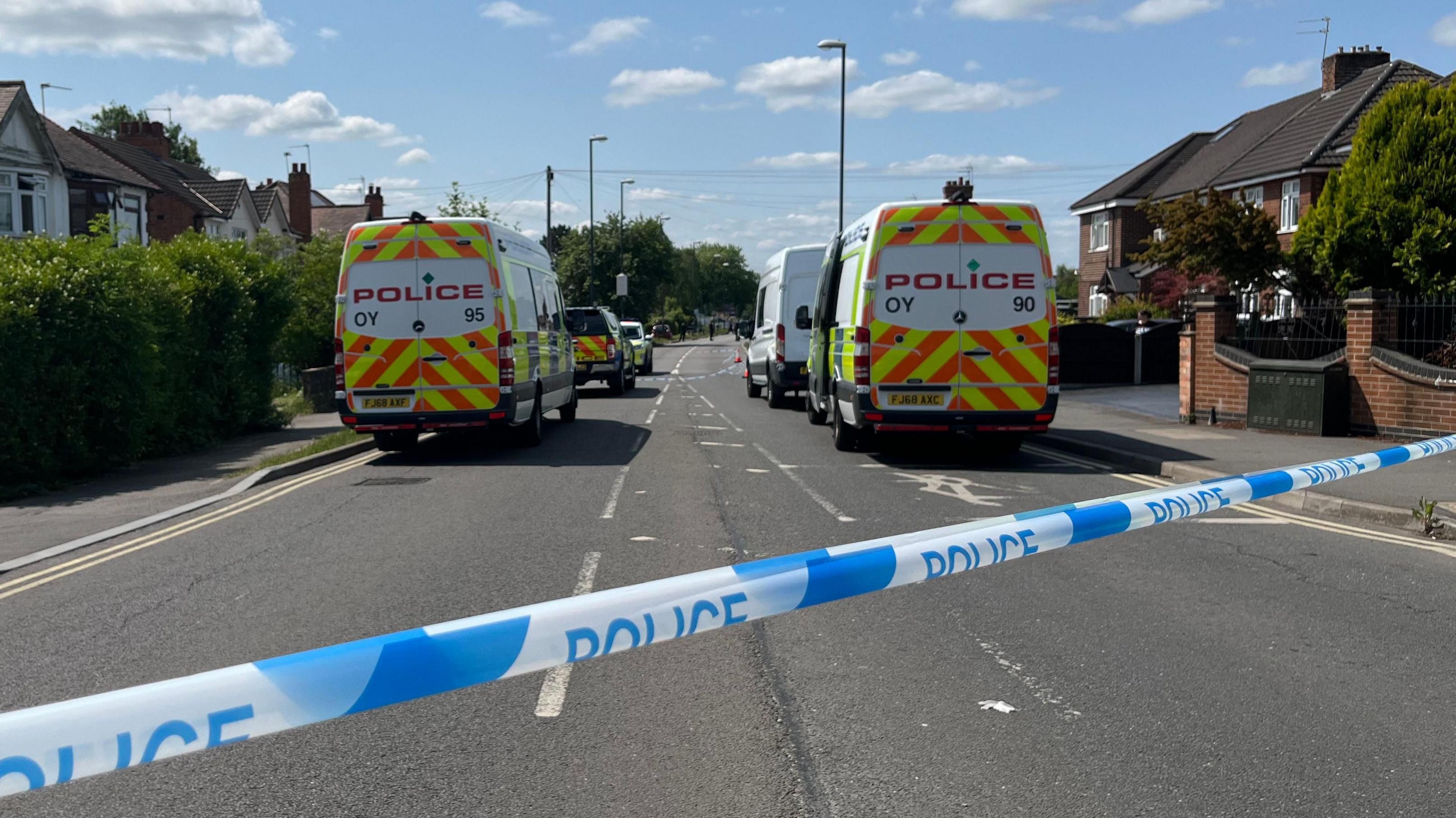 A police cordon tape across a residential street with police vans in the background