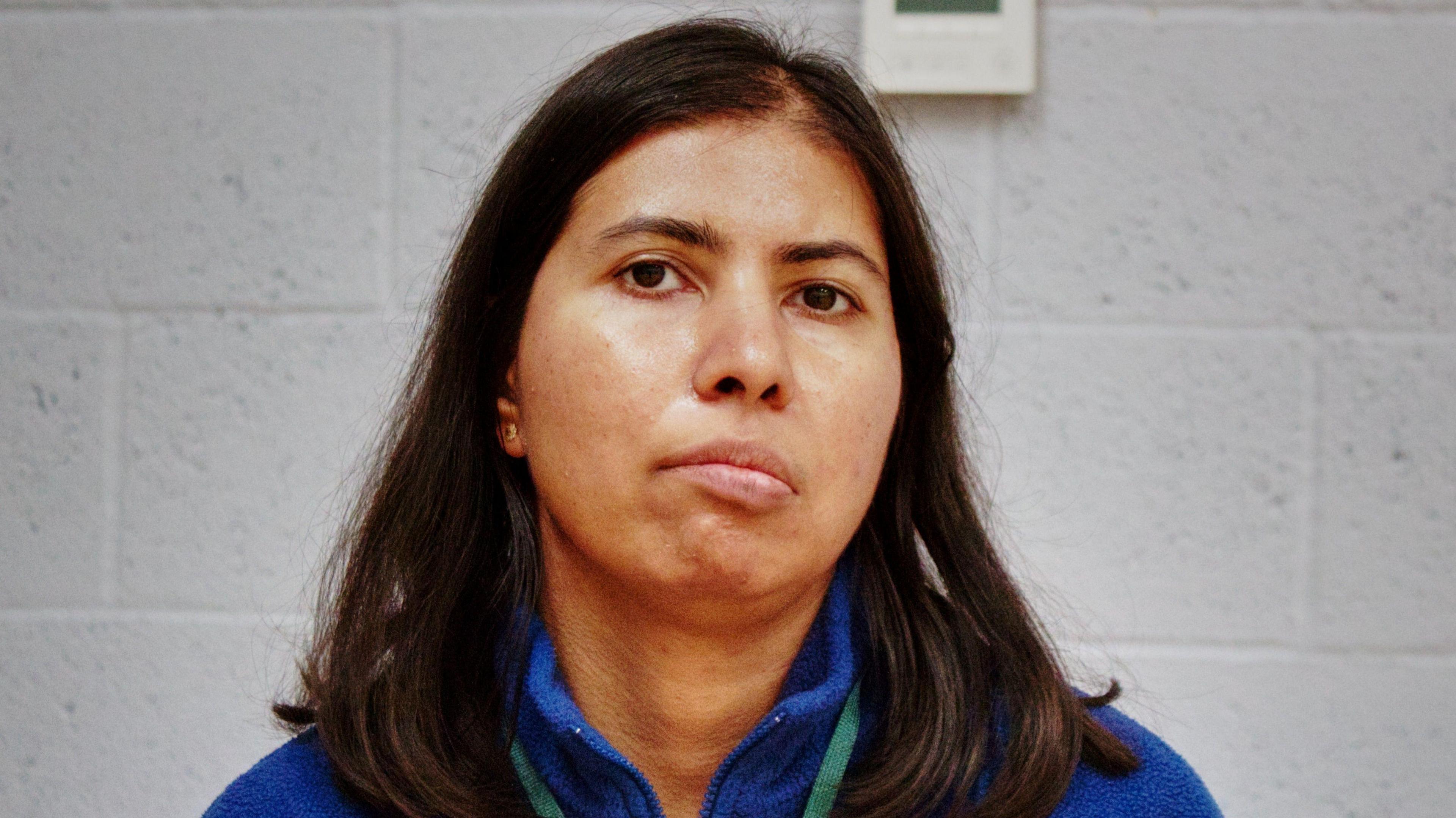 Janisha, looking at the camera, wearing a blue fleece top, with a green lanyard round her neck. She has long dark hair, which is down, and is standing in front of a grey wall. 