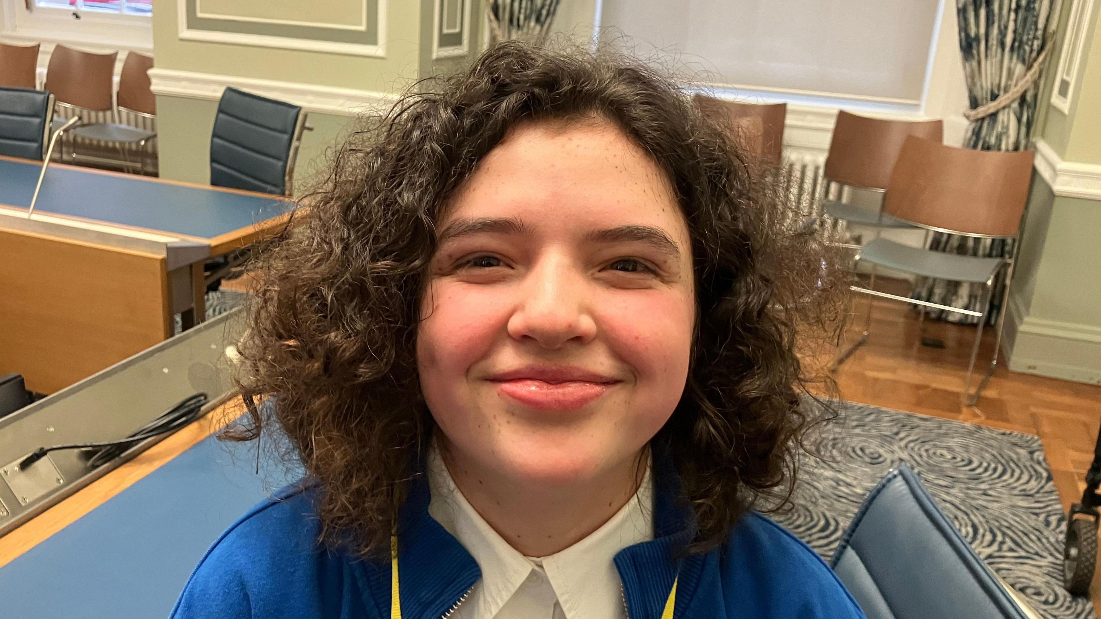 Ellie-May has short dark curly hair. She is smiling in the picture, and is wearing a white shirt and blue cardigan.