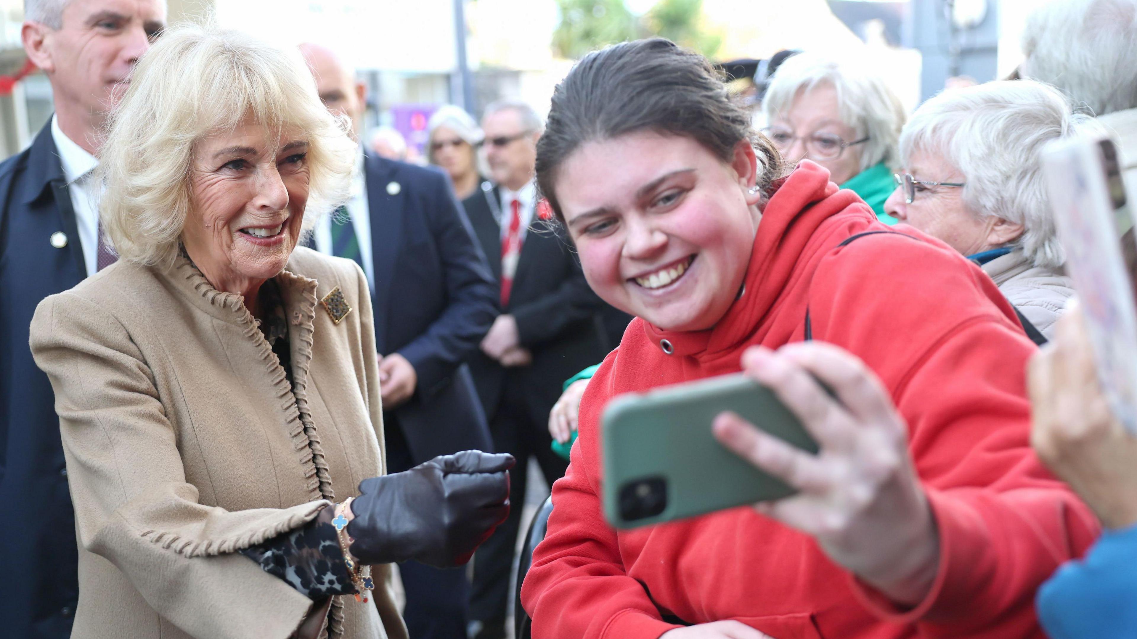 A woman in a red jumper holds a green phone and takes a selfie with Camilla