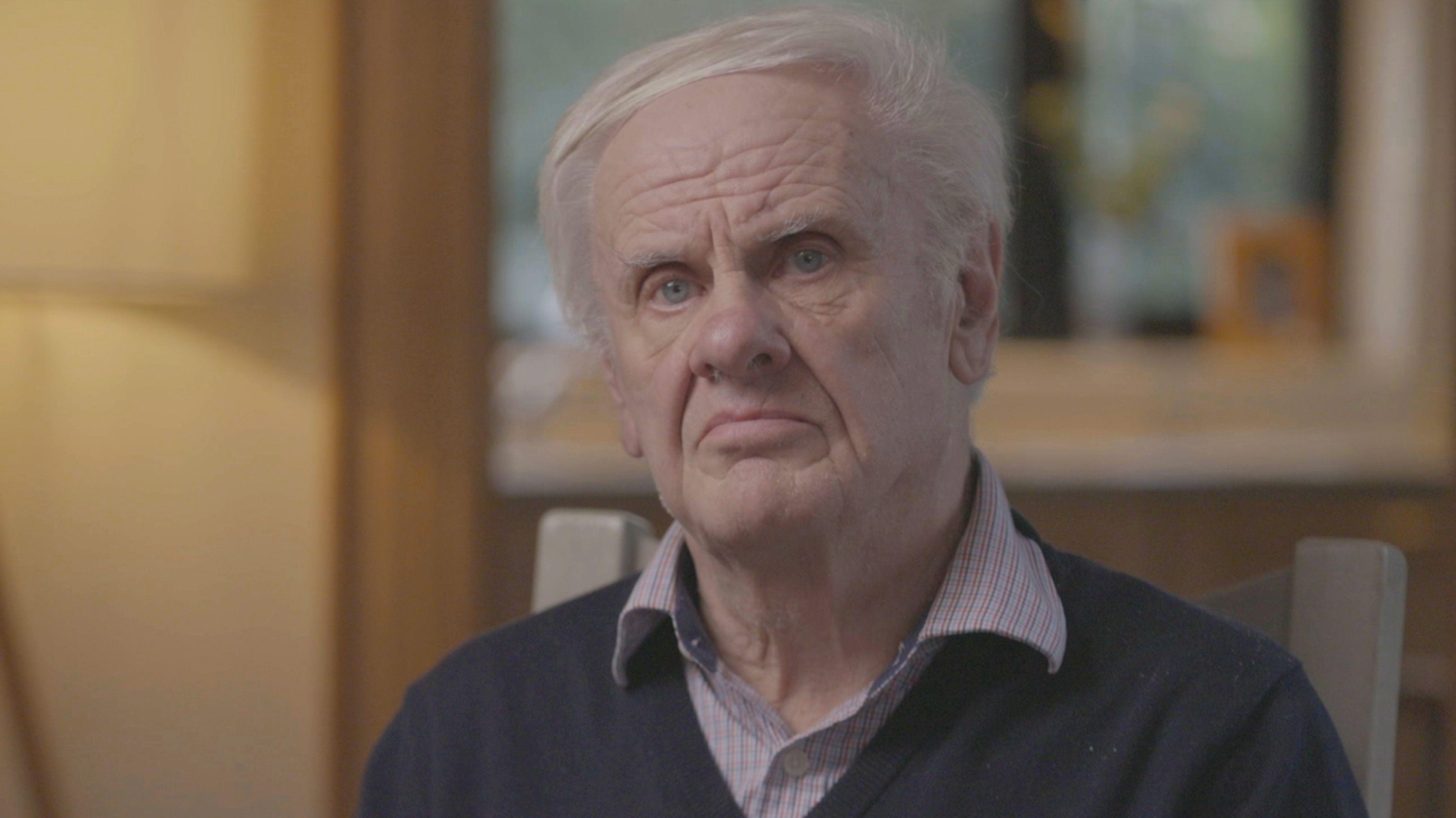 A man with grey, thinning hair looks at the camera. His hair is swept to the side as he sits on a wooden backed chair. The man is wearing a dark jumper and chequered shirt, with an open collar. Behind him is a blurred view of a window or bright painting, surrounded by yellow walls.