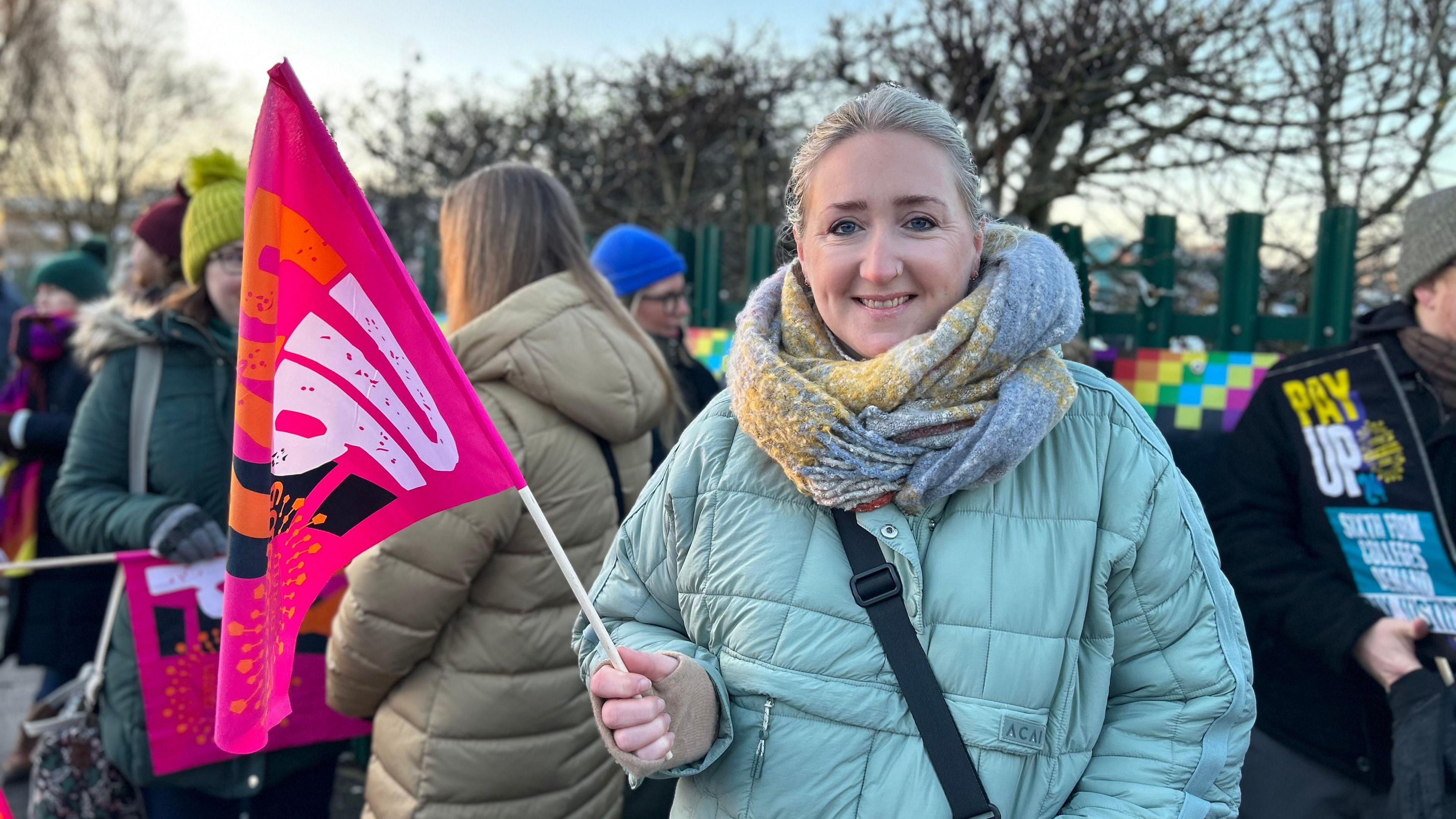 A woman holding a pink NEU flag outside the college gates. She is wearing a light-blue padded coat and a large grey and yellow scarf. Fellow picketers can be seen behind her.