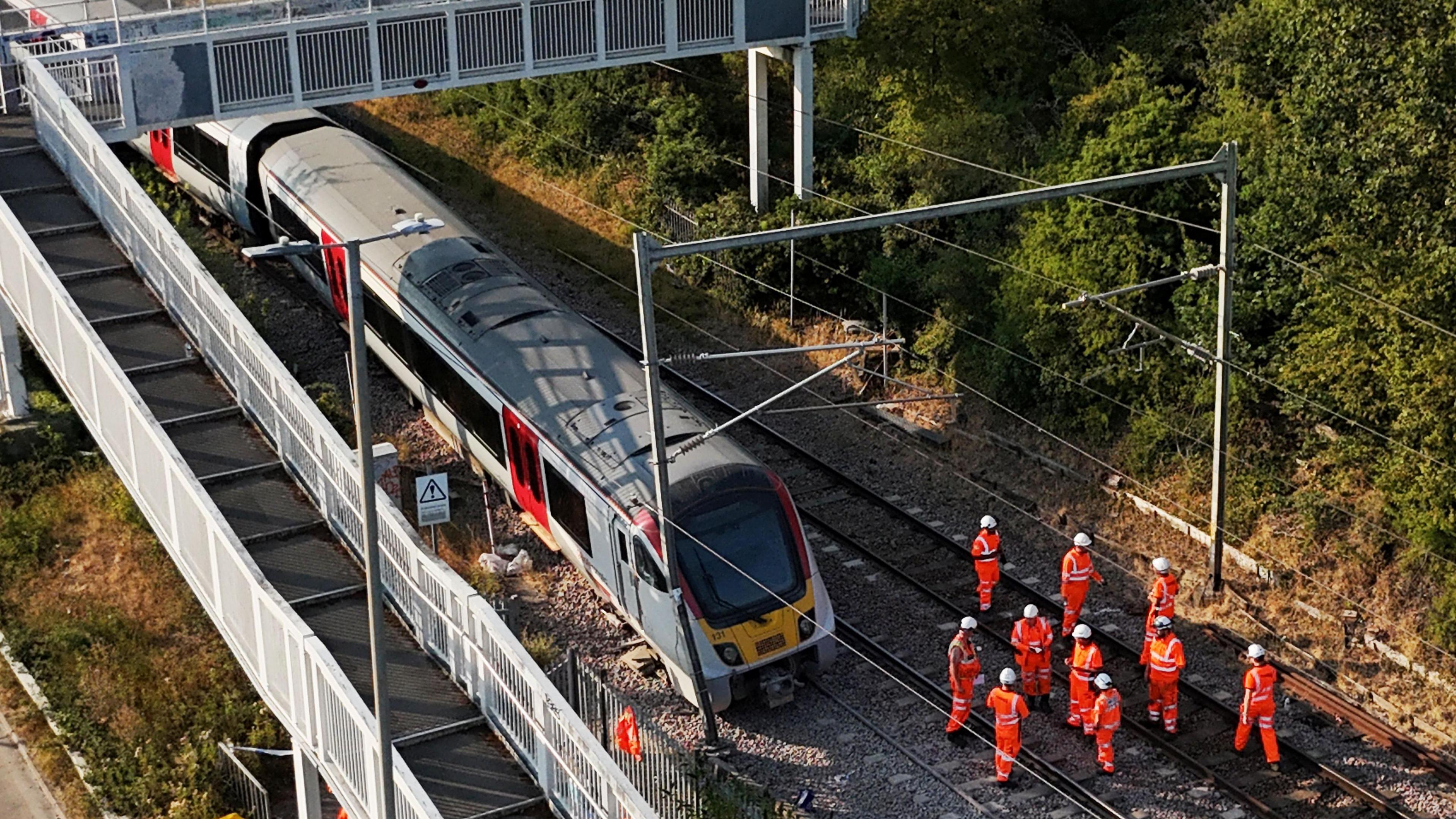 A photograph of the Greater Anglia train with part of the train off the track at Wickford in Essex. The train has been stopped and is not moving. Around 10 or more people are wearing orange fluorescent hi-vis with white helmets and are stood on the tracks to the right of the train.