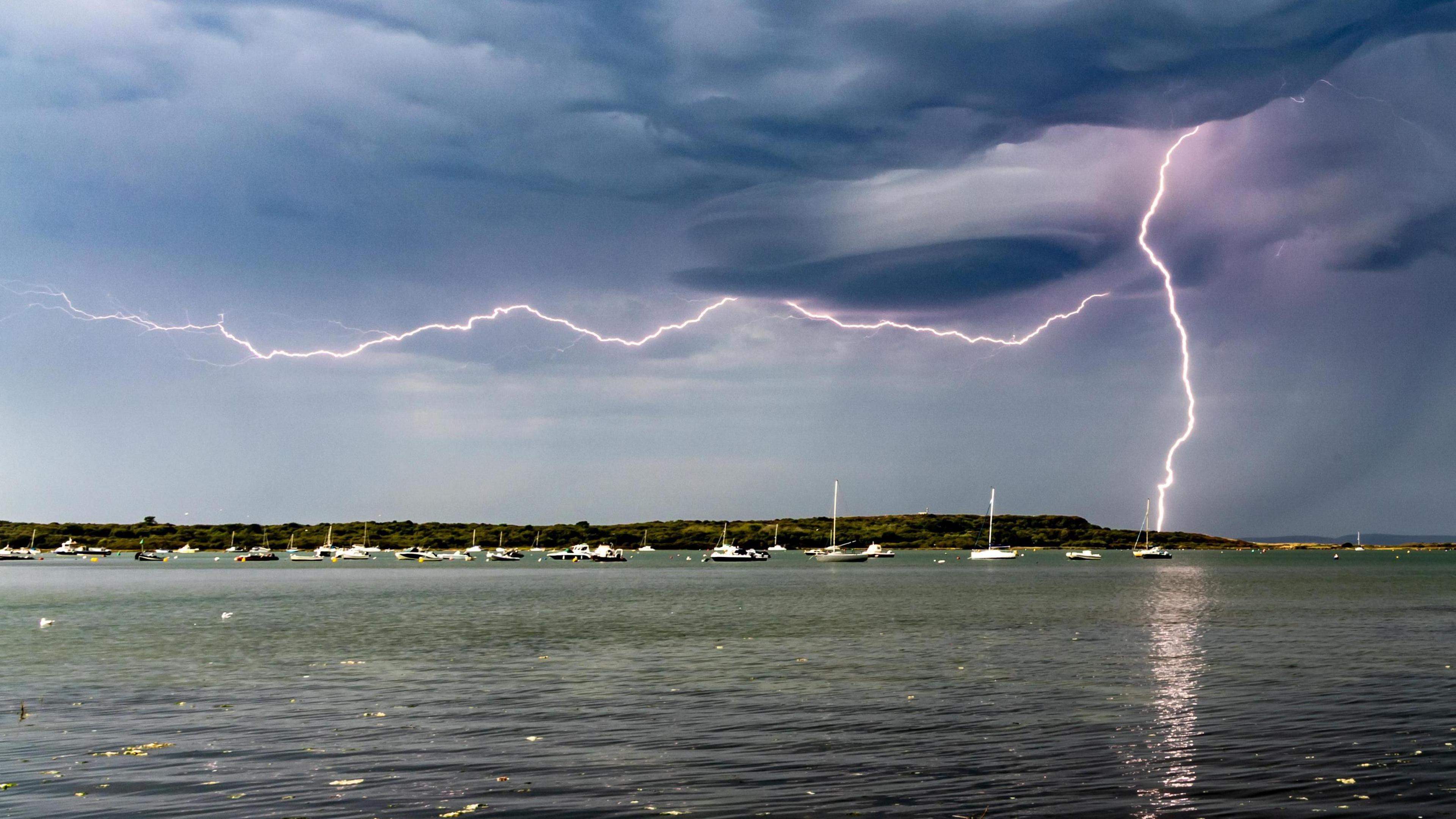 A seascape with small boats in the distance and sand dunes on the horizon. The evening skies are full of dark clouds and a lightning strike is angling to the ground and across the clouds simultaneously.