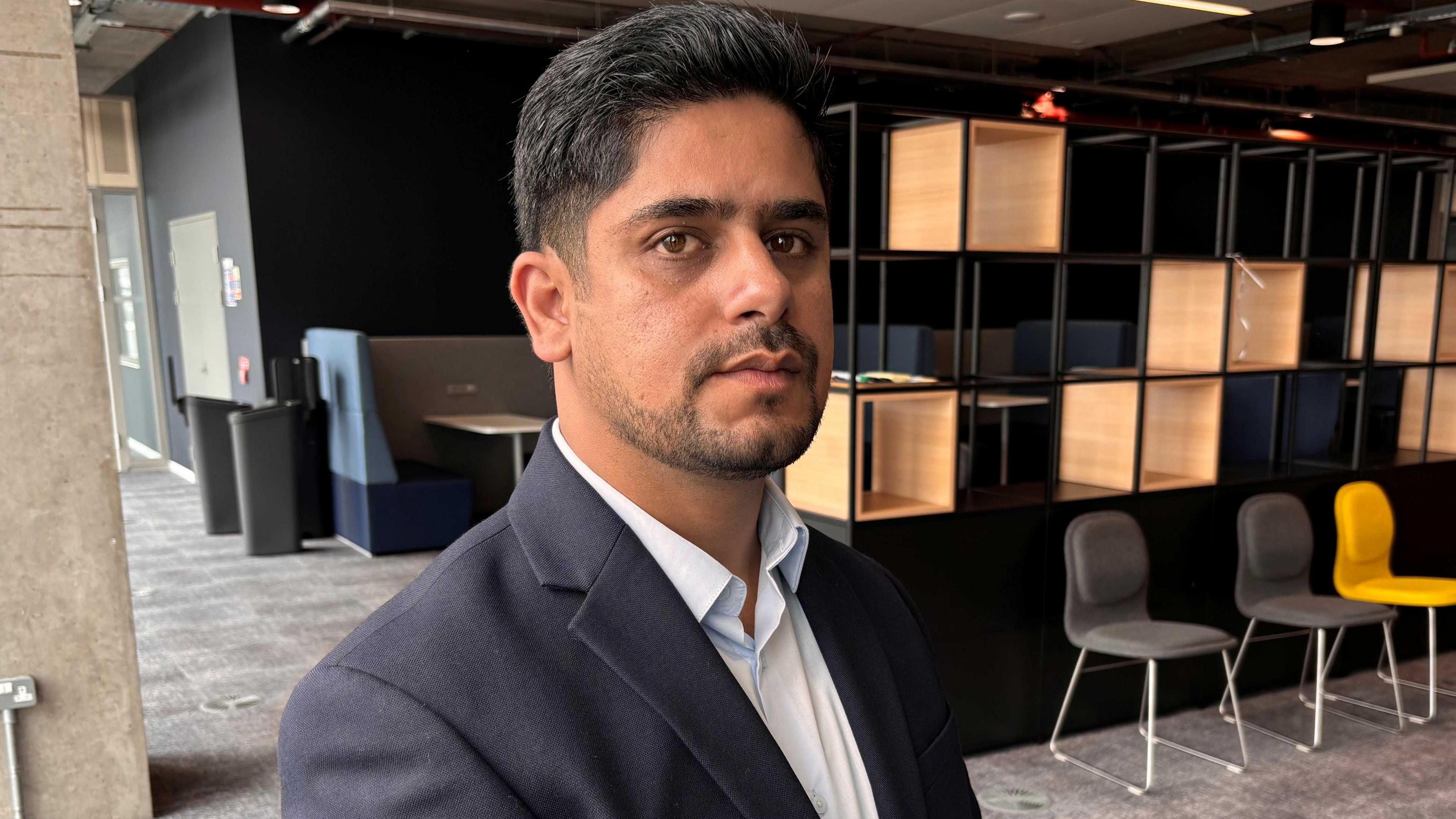 A man with black hair and stubble, wearing a dark blazer and light blue shirt, stares into the camera with a neutral expression. He is in an office and is standing in front of a row of shelves and some desk chairs.