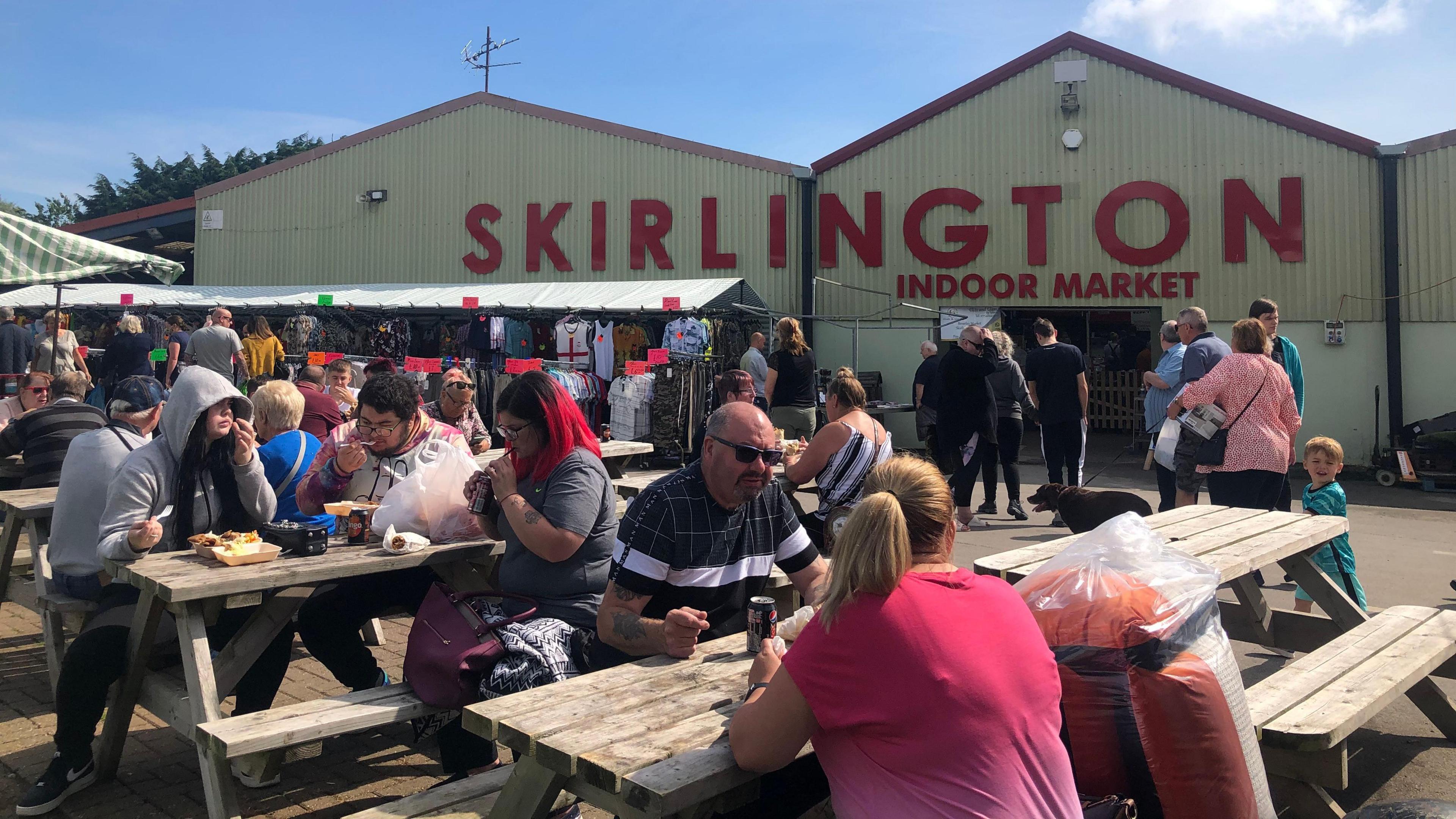 Several benches located in an outside seating area with several people sat on them with canned drinks and food. In the background is a warehouse-type building which says 'Skirlington Indoor Market' in large red letters.