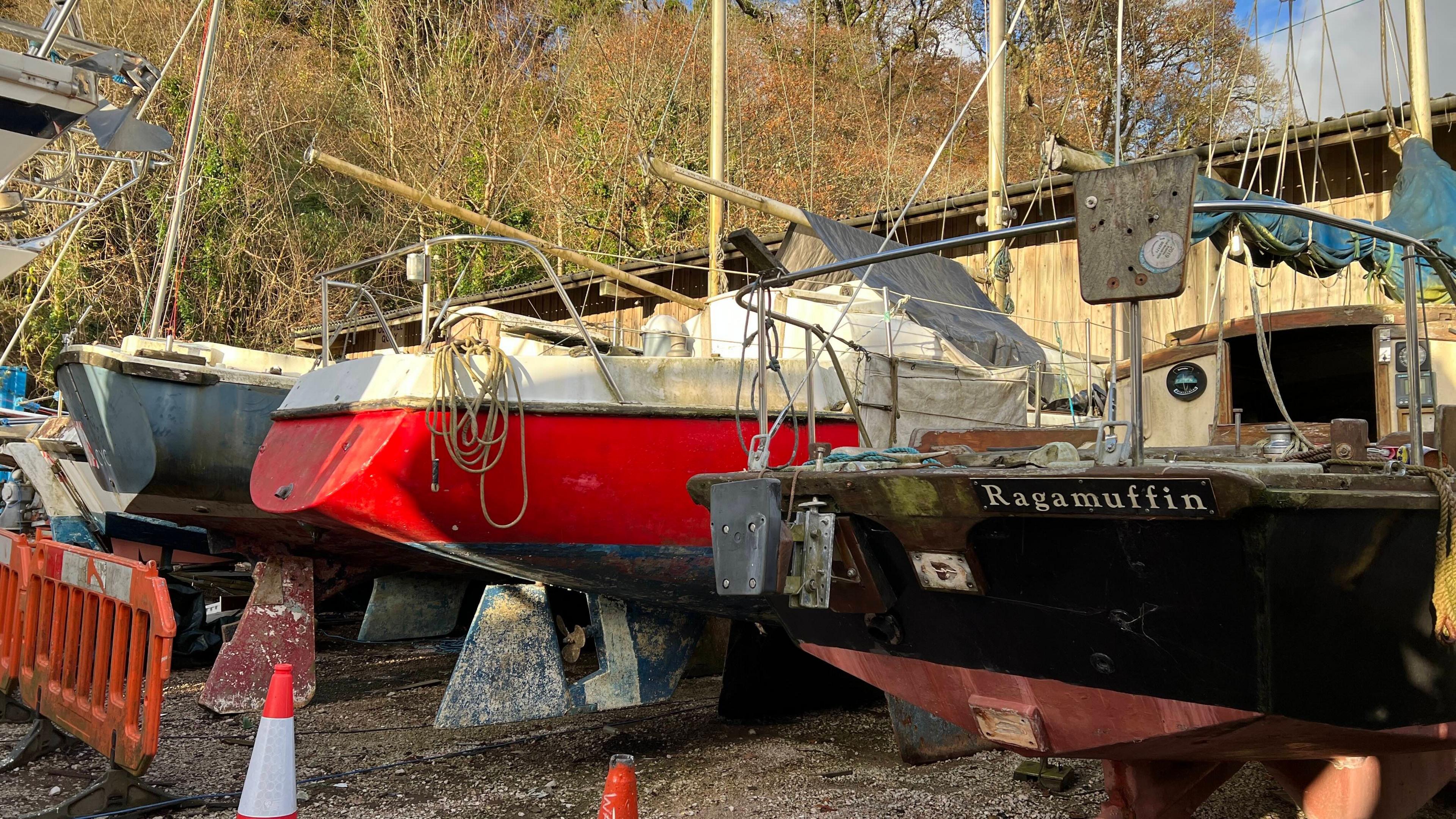 A row of three old boats, one black, one red and another grey with masts in a boatyard.