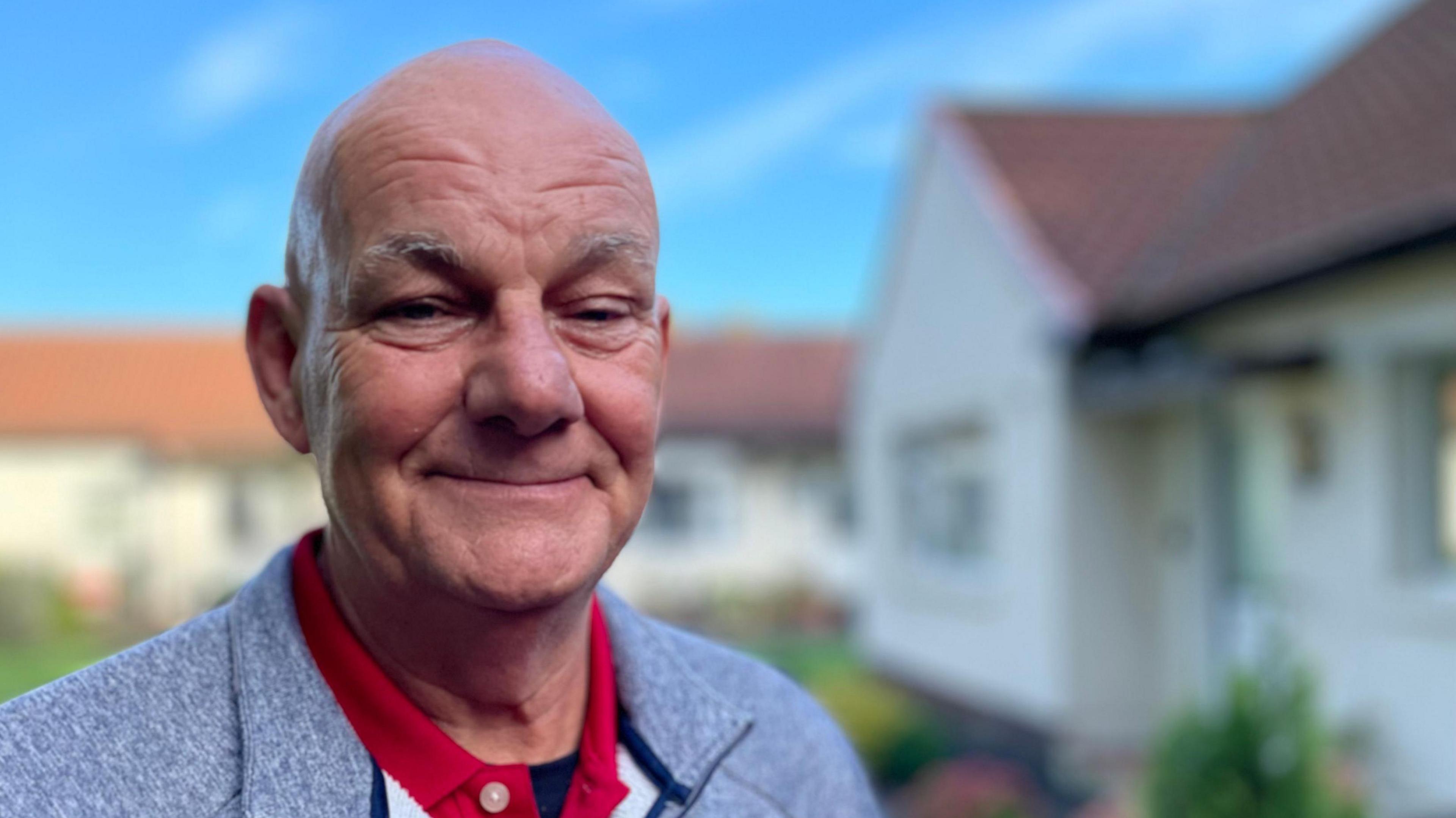 Paul McBean smiles with closed lips as he stands under a blue sky in front of rows of white bungalows. He wears a red polo shirt and a light grey jacket.