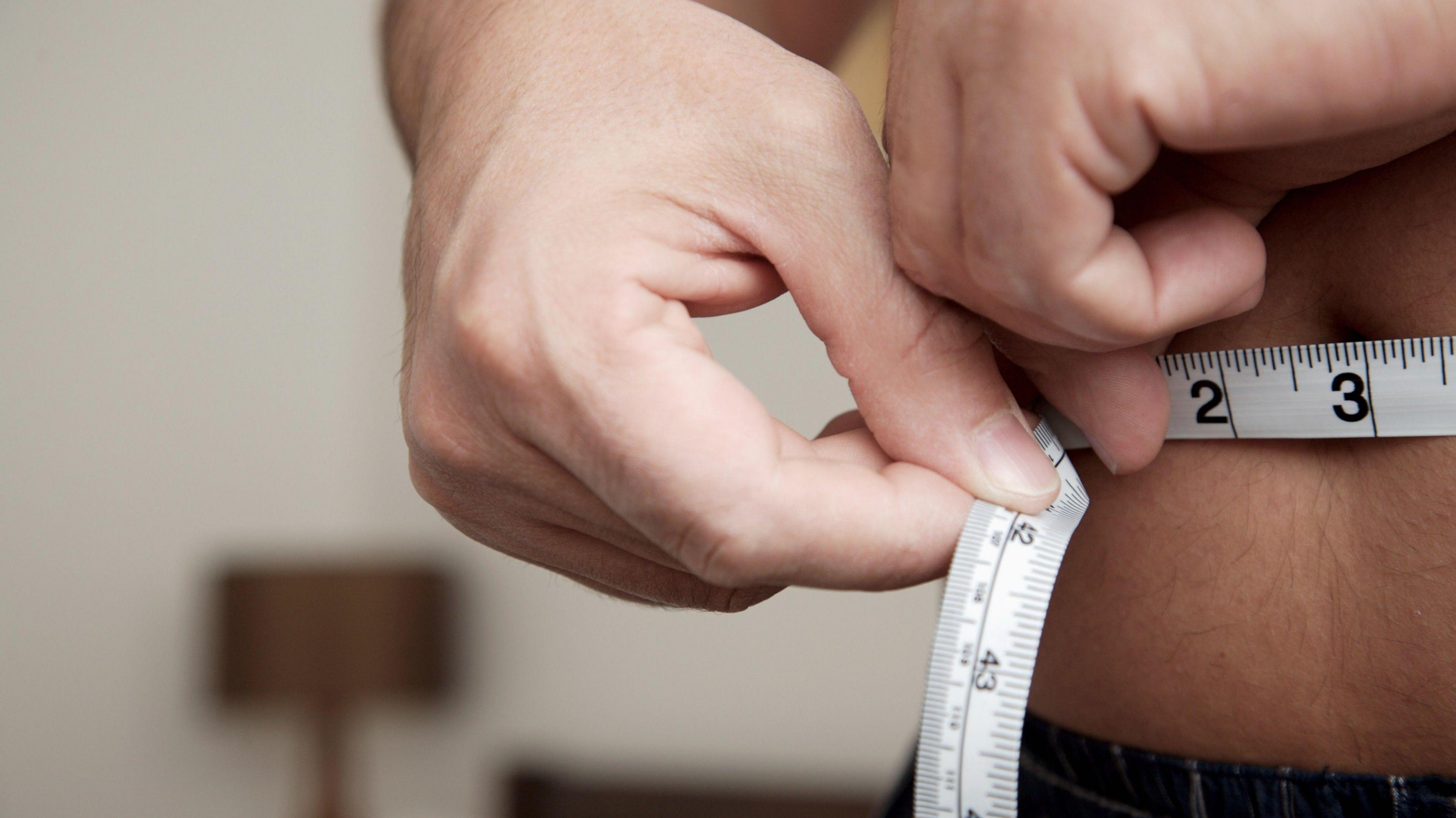 A close up of a man measuring his waist with a tape measure.