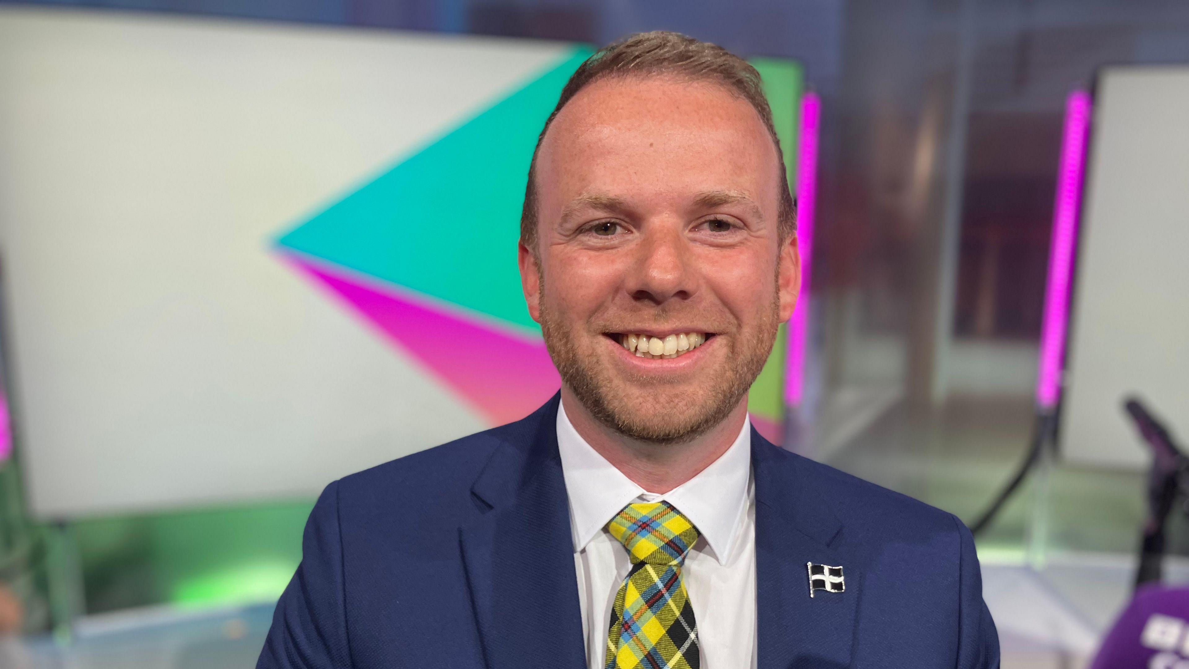Ben Maguire is smiling broadly at the camera. He is wearing a white shirt, blue jacket and a Cornish tartan tie. He has short blond hair and a slight beard. There is St Piran flag badge in the button hole of his jacket. Behind him are neon-lit panels.