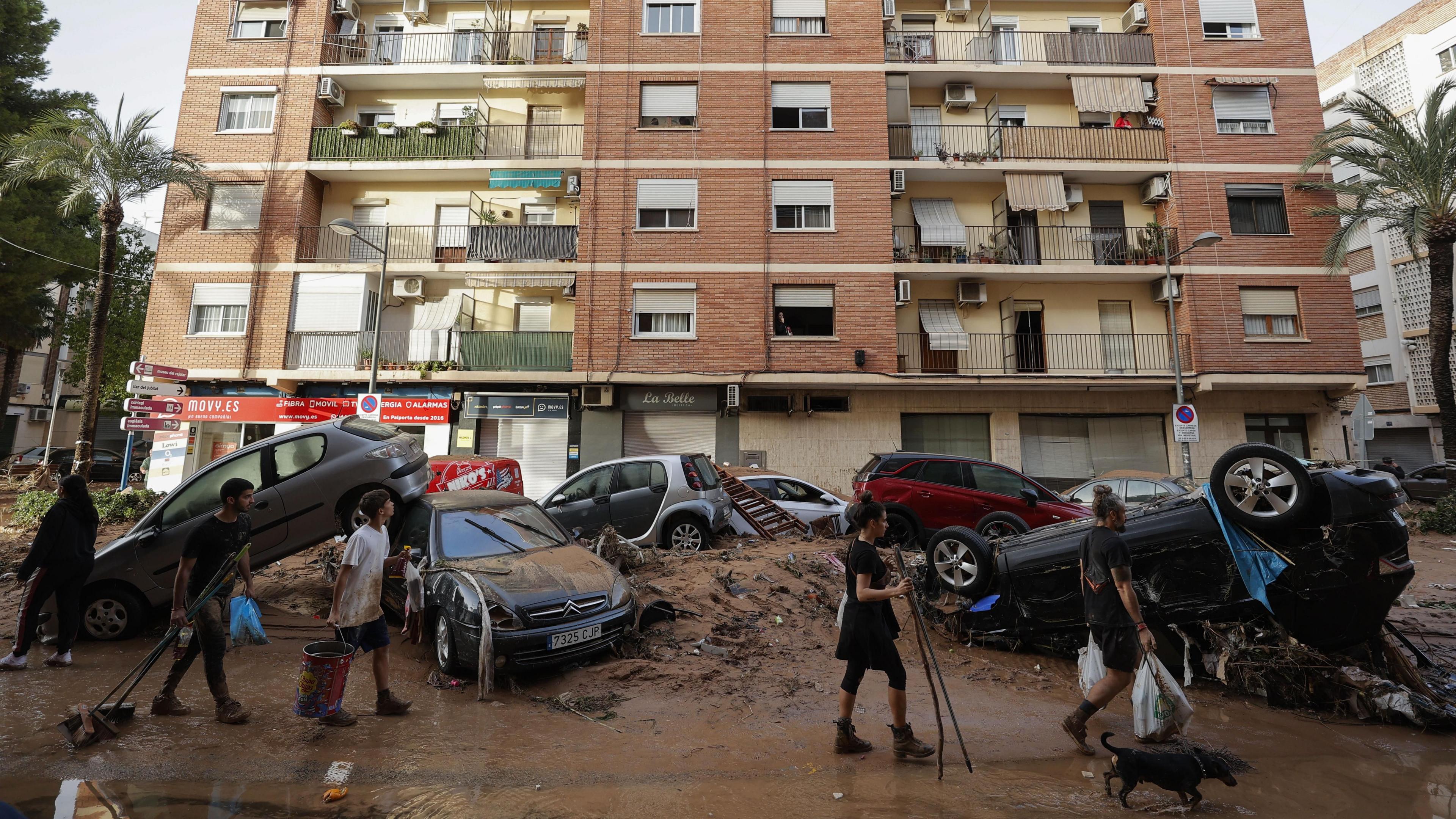 People carrying buckets and brooms walk past piled-up cars along a mud-covered street