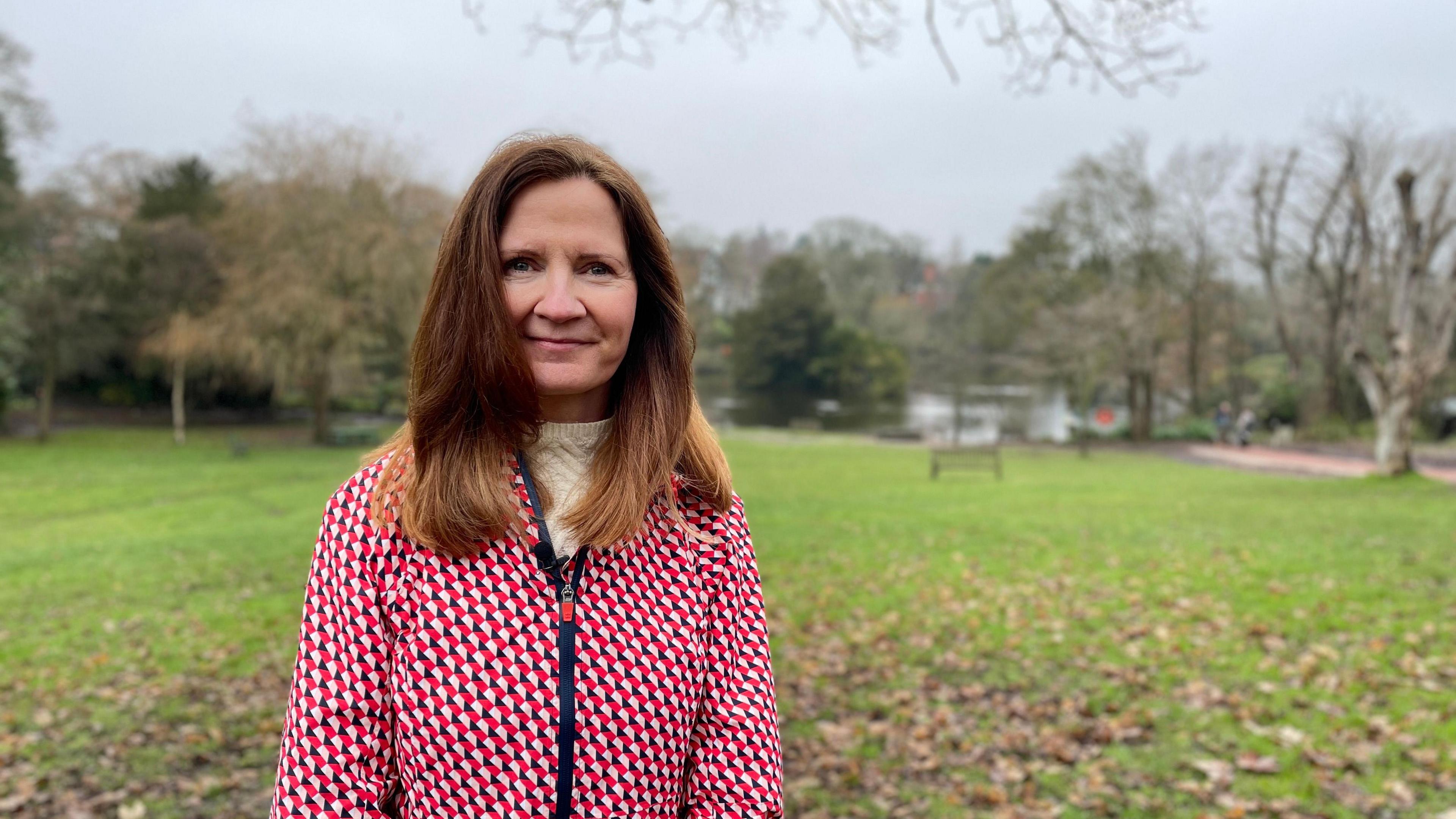 Kirsten de Vos at Moseley Park in Birmingham. She has long, light brown hair, past her shoulders and wears a red and pink patterned coat over a white jumper. Behind her is green grass with brown leaves on it and, further back, a bench with trees in the distance.