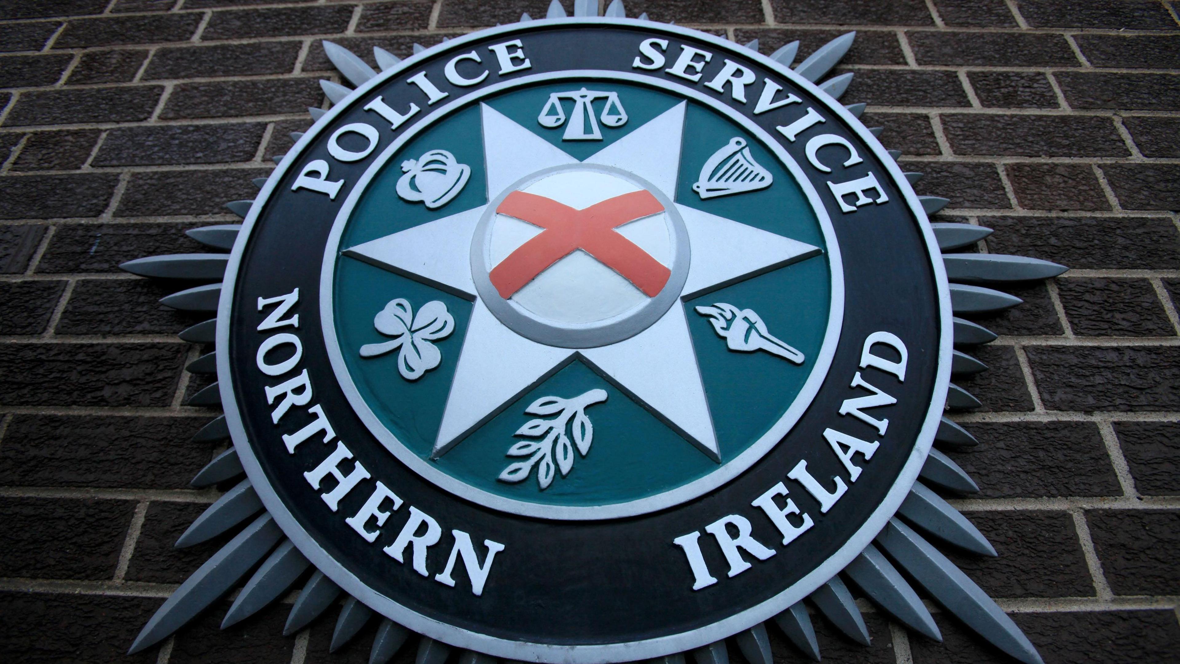 The PSNI emblem on a brick wall. It includes scales, a harp, a torch, a leaf, a shamrock and a crown.
