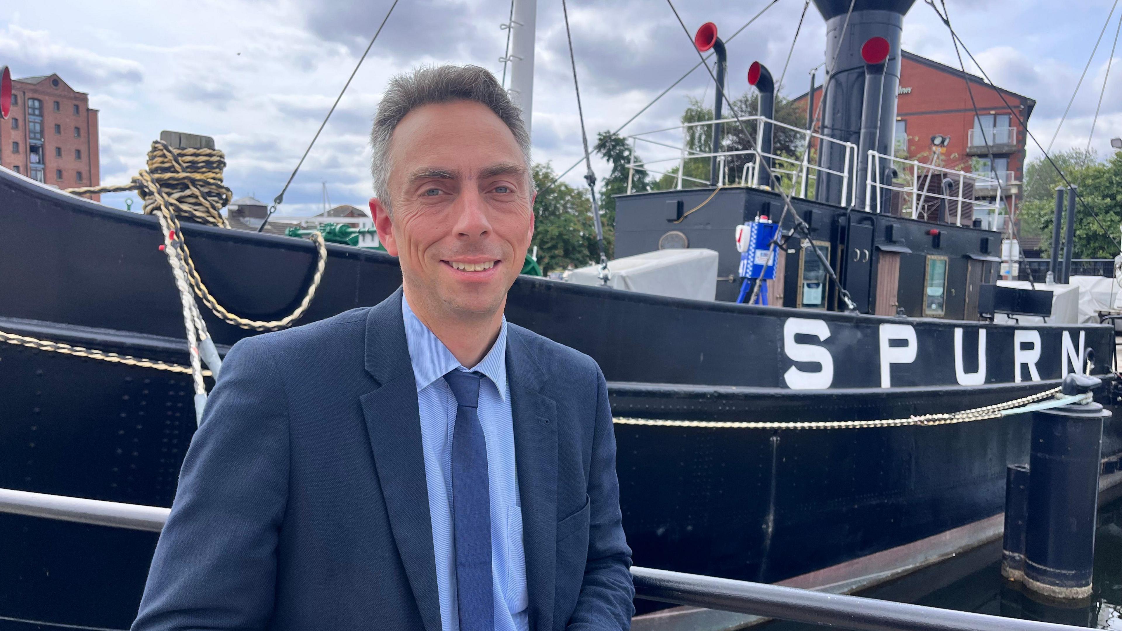 Mike Ross, the leader of Hull City Council stands in front of the Spurn Lightship which is berthed in Hull marina and looks directly at the camera wearing a navy blue suit and tie.