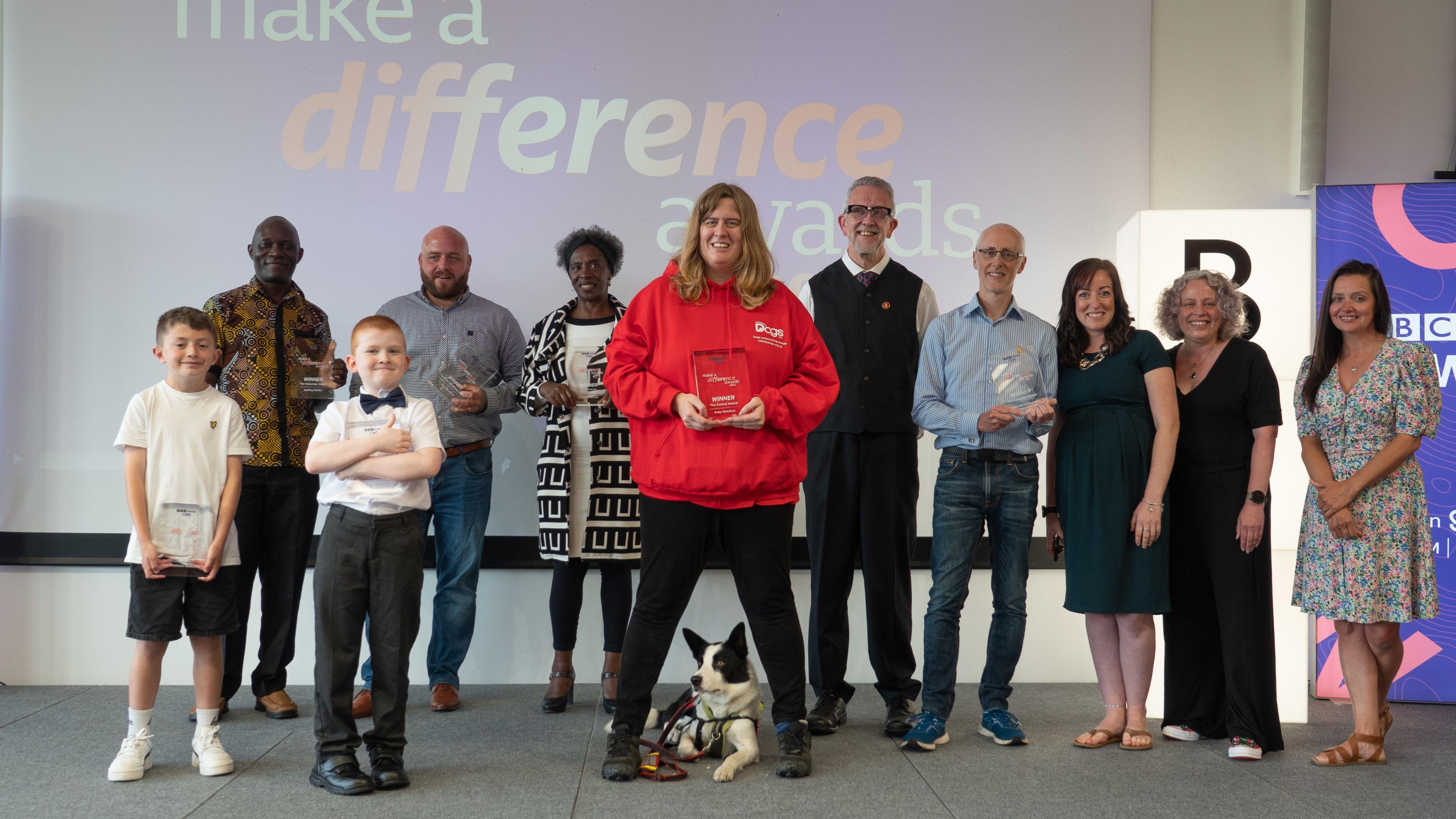 11 people stand on a stage. Some of them are holding glass awards which they have won