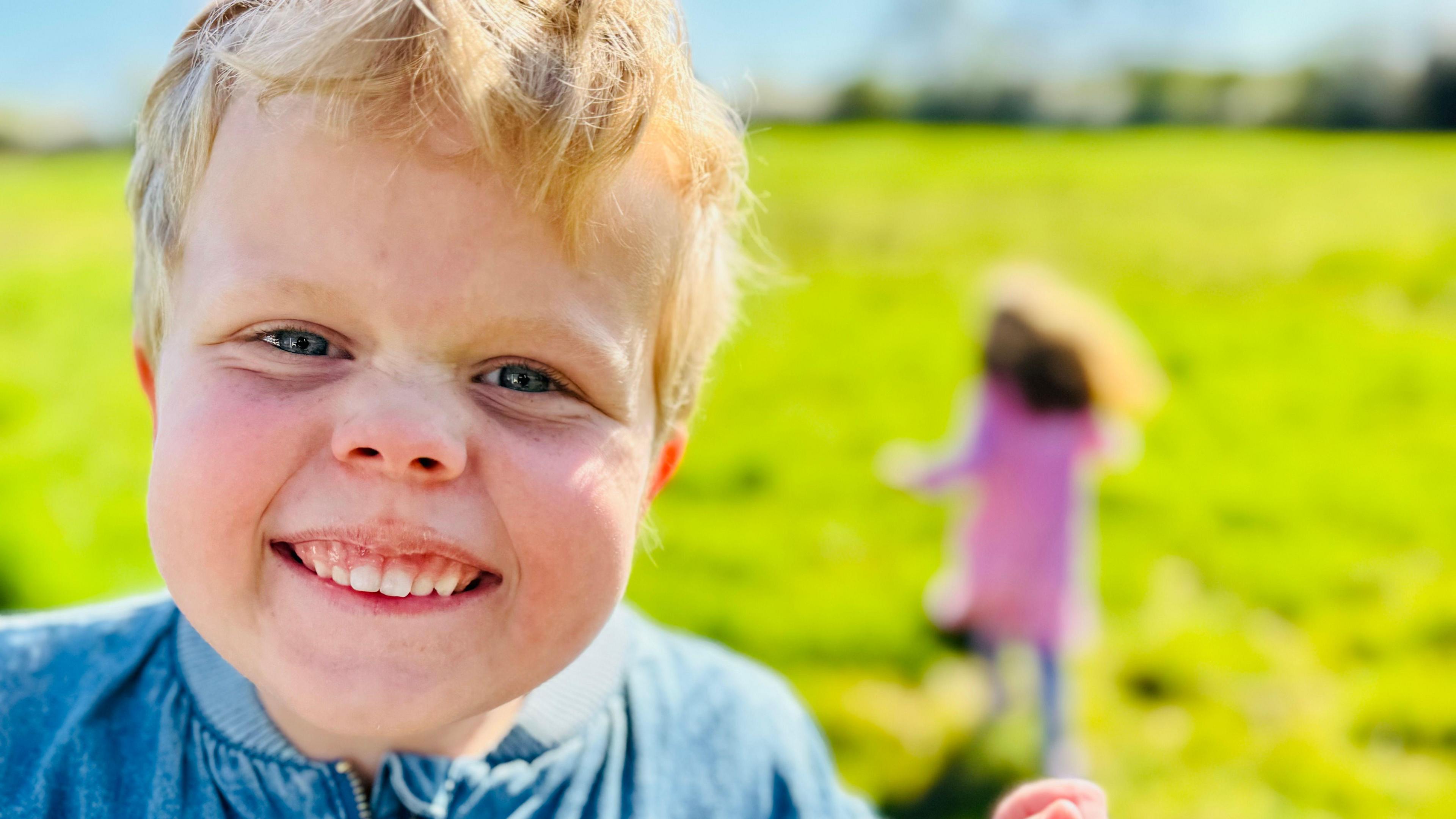 Octavia smiling at the camera wearing a blue top. Her sister can be seen out of focus in the background in a pink dress. They are surrounded by grass.