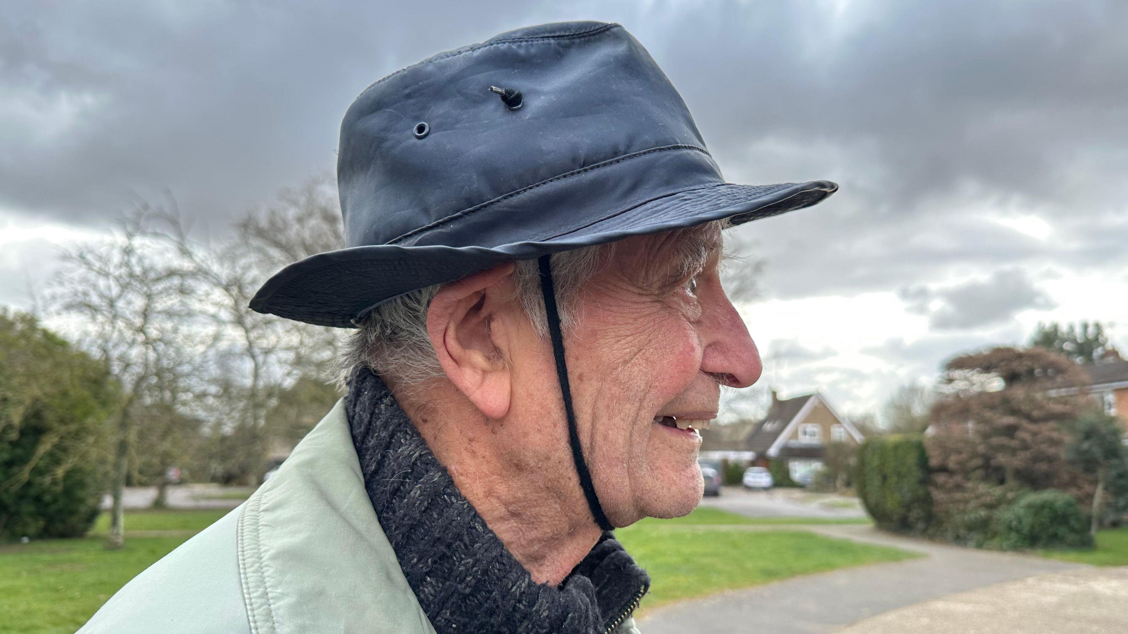 91-year old Glyn Parry is stood outside facing side-on to the camera. He is wearing a hat with a piece of shoelace tied underneath his chin to keep his hat on.