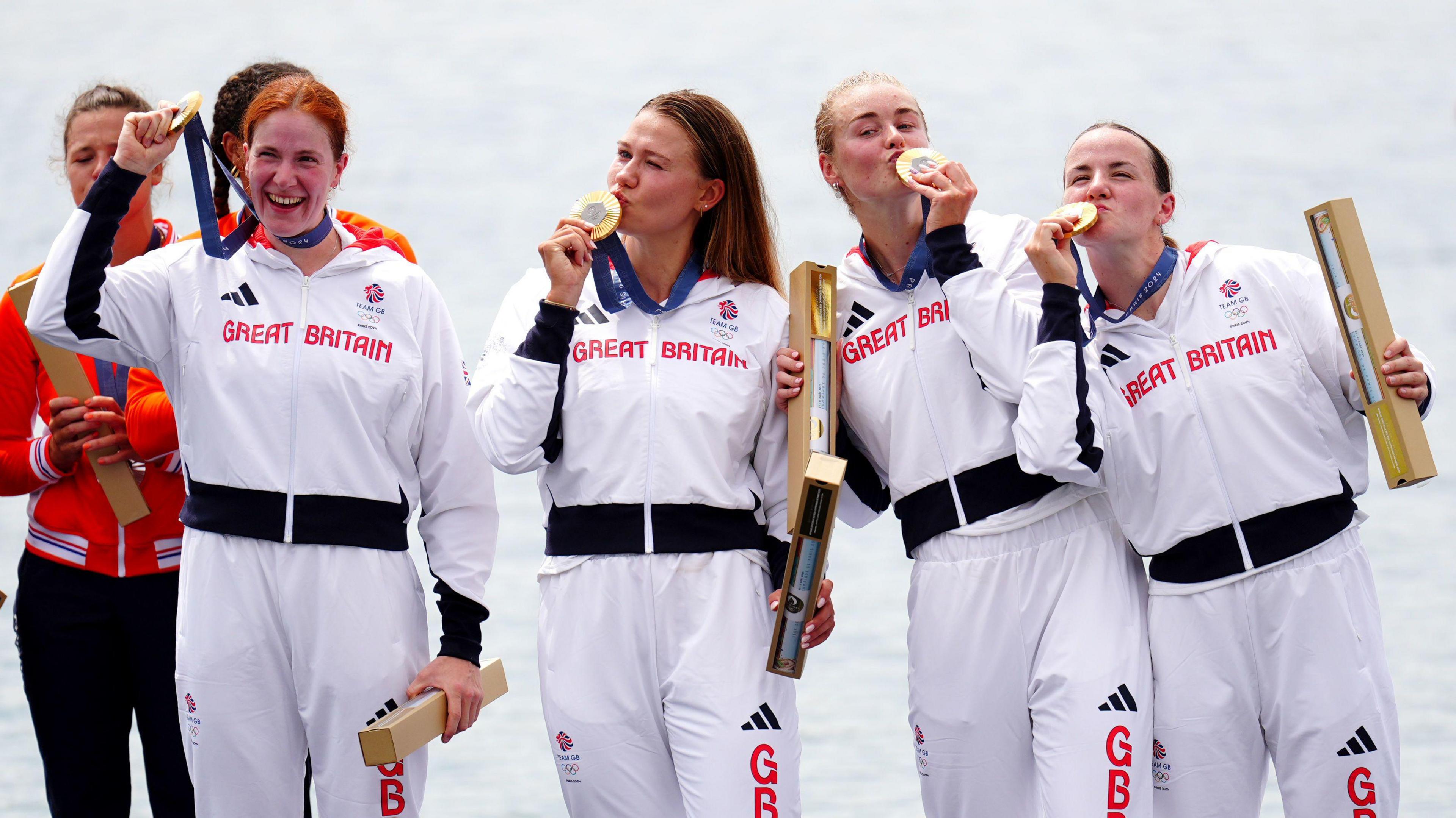 four women are stood on a platform wearing a white tracksuit with the red great Britain Olympic branding on their tops. They are all looking in random directions, Kissing their gold medals and holding a gold tube of a map in their opposite hands. 