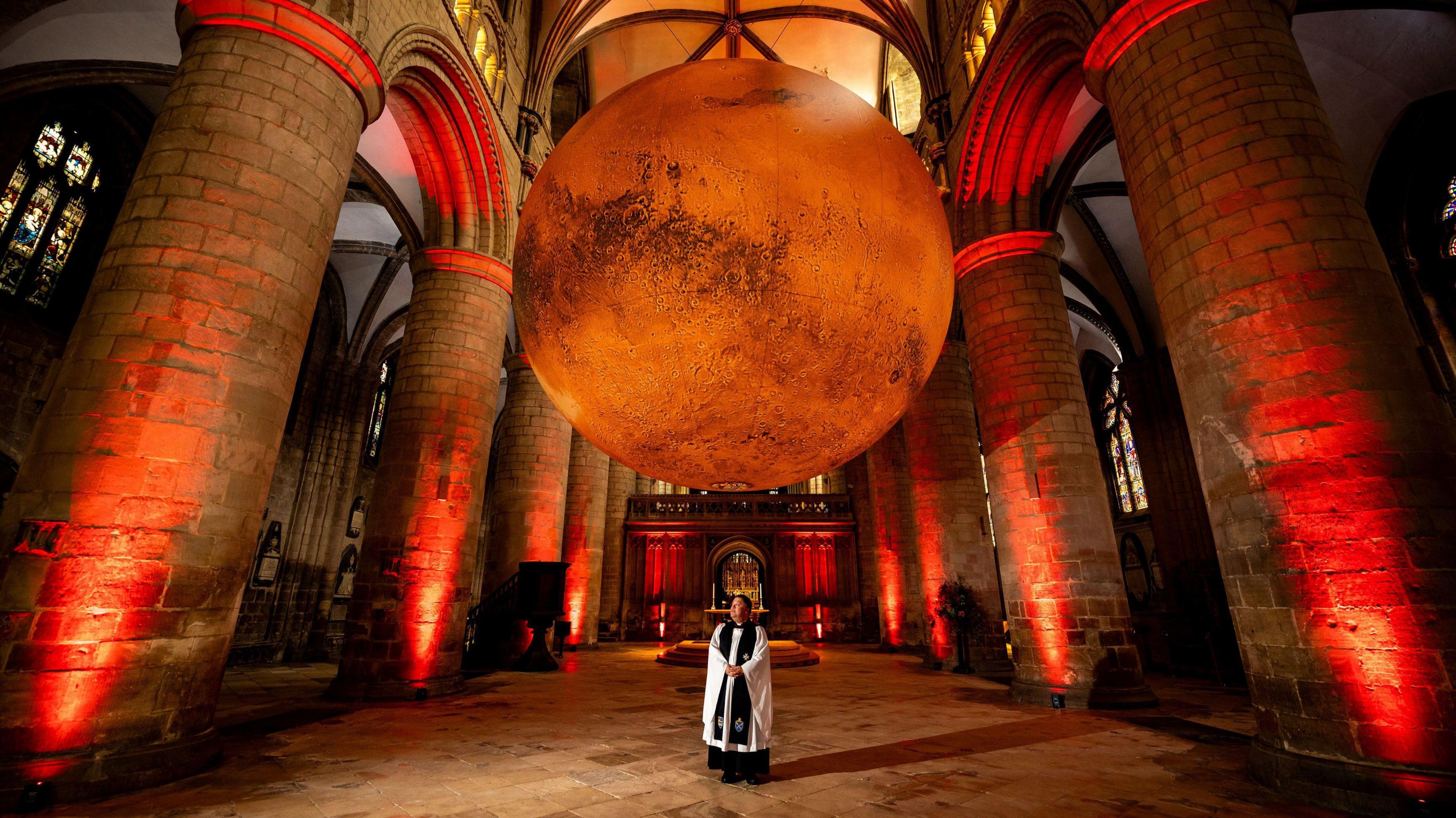 The huge Mars sculpture hanging in Gloucester Cathedral with a member of the clergy standing underneath it. The cathedral space is lit up by red uplighters