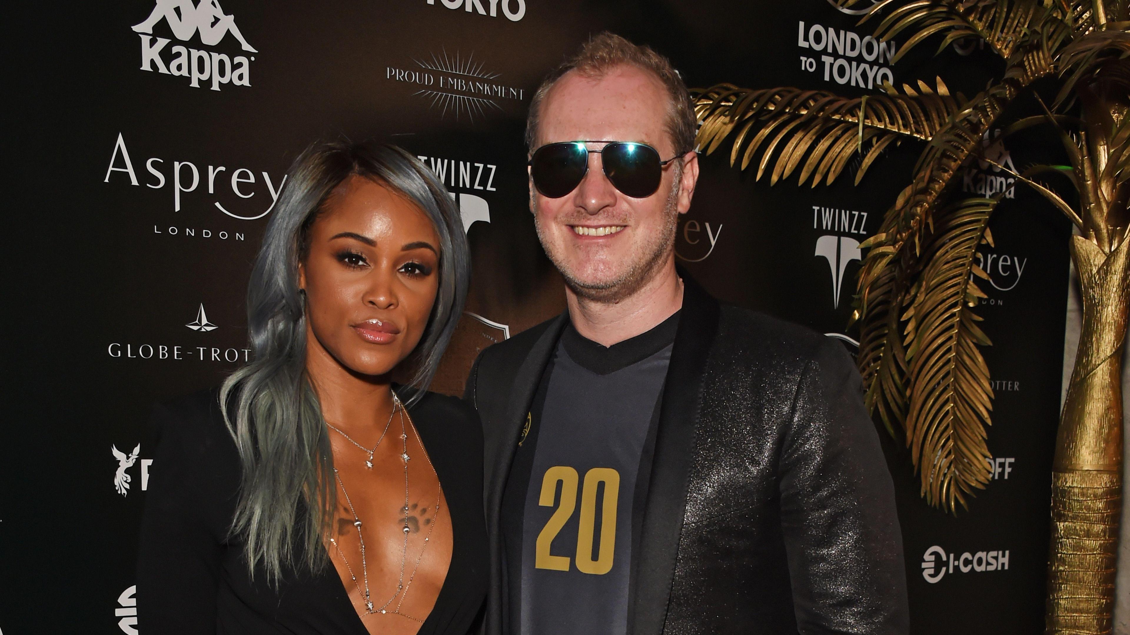 A woman with light grey is stood with a man in a sparkly jacket and a grey t-shirt with a gold 20 on the front. He is also wearing large sunglasses next to a gold tree.